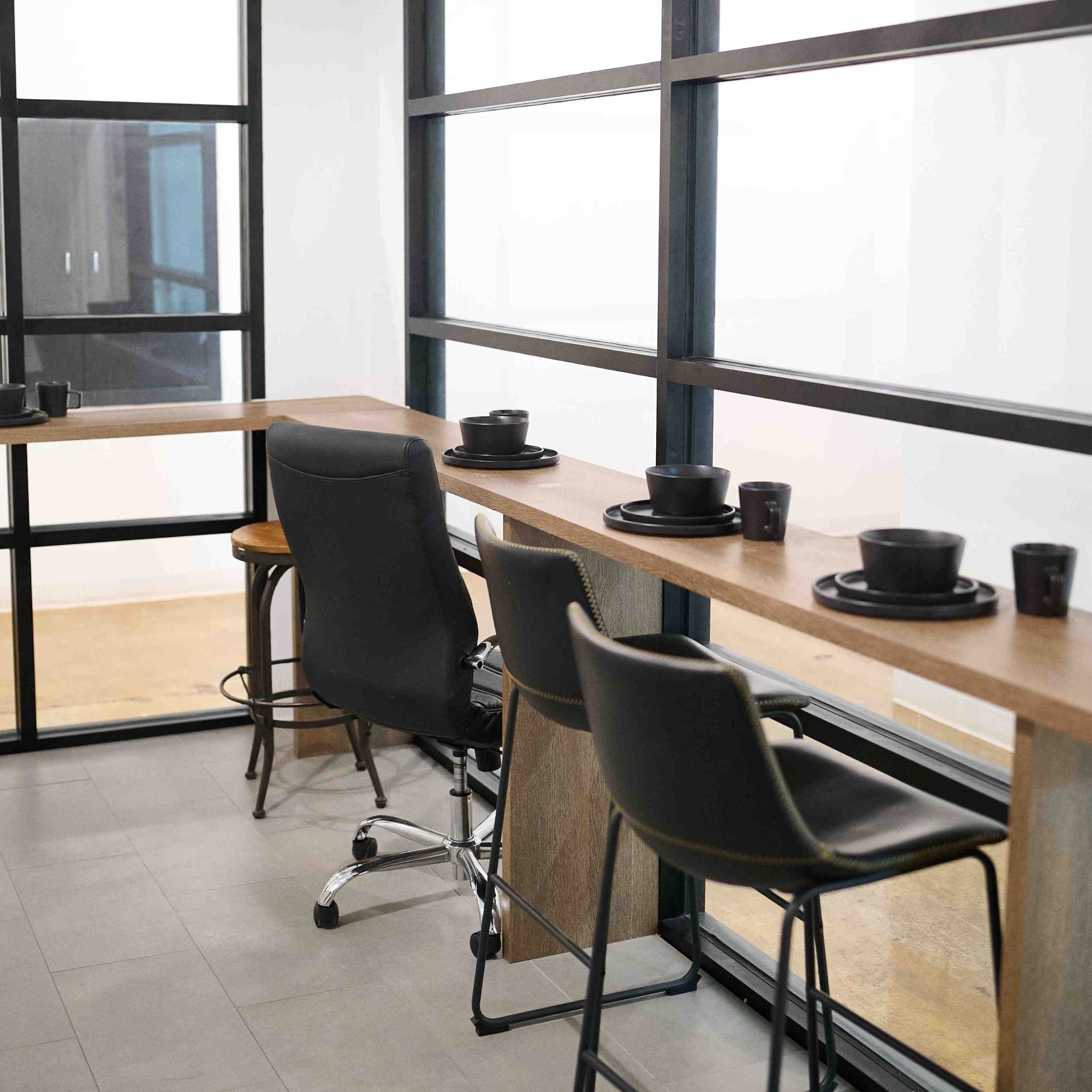 A modern workspace with a long wooden counter, black chairs, and tableware, situated near large glass windows.