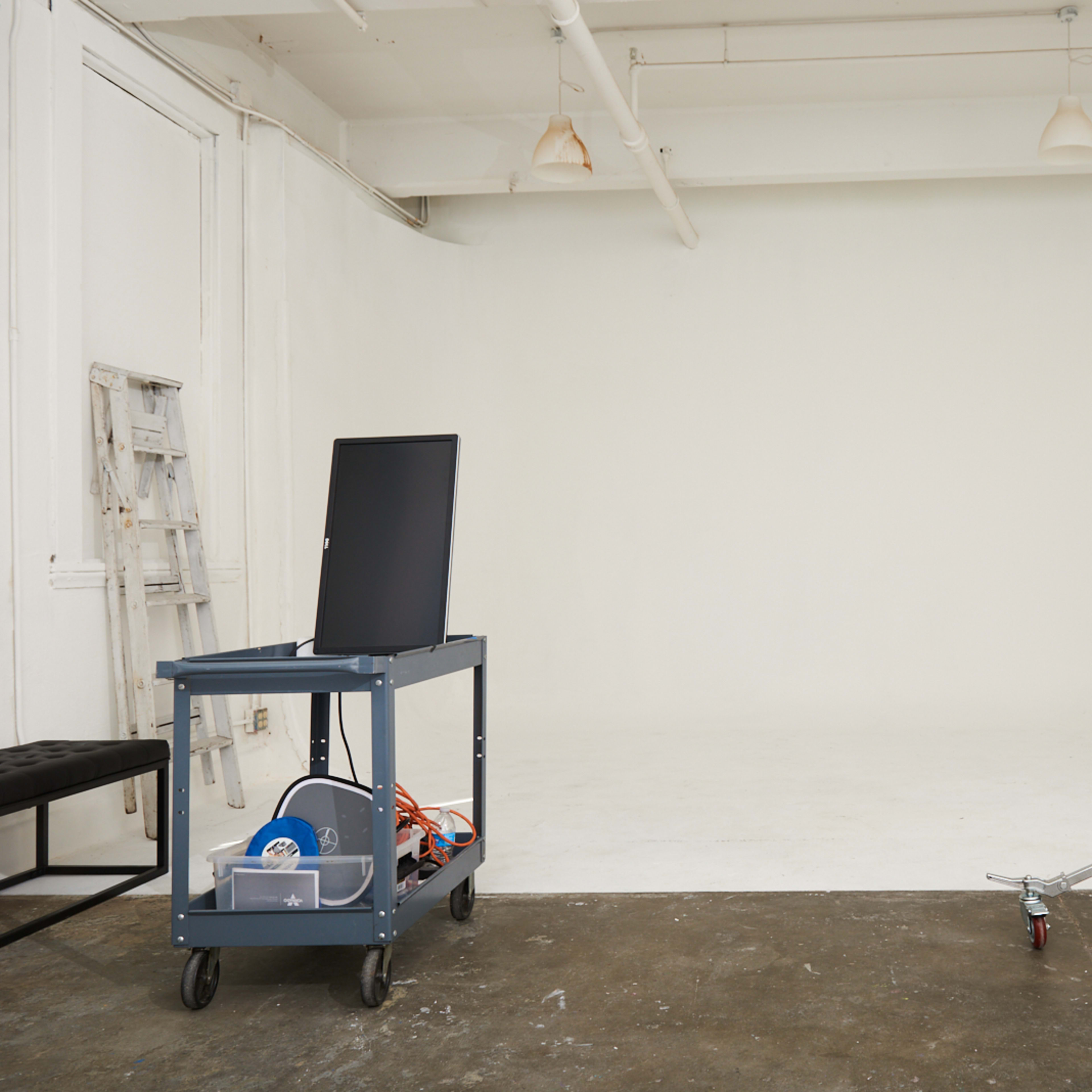 A modern studio space features a blank white wall, a cart with equipment, a light stand, and a chair in the corner.