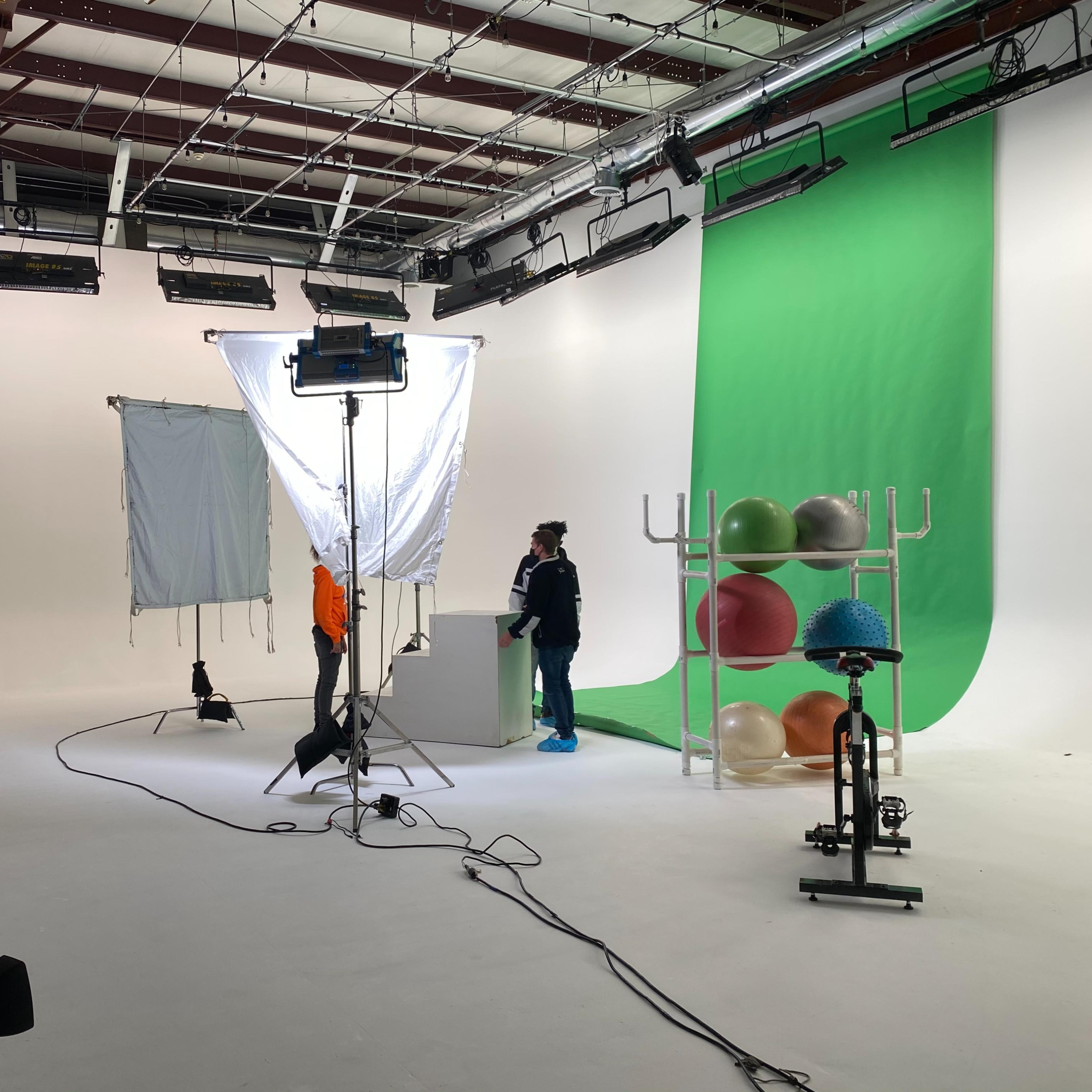 A photography studio is set up with a green backdrop, lighting equipment, and various props, including exercise balls and a stationary bike.