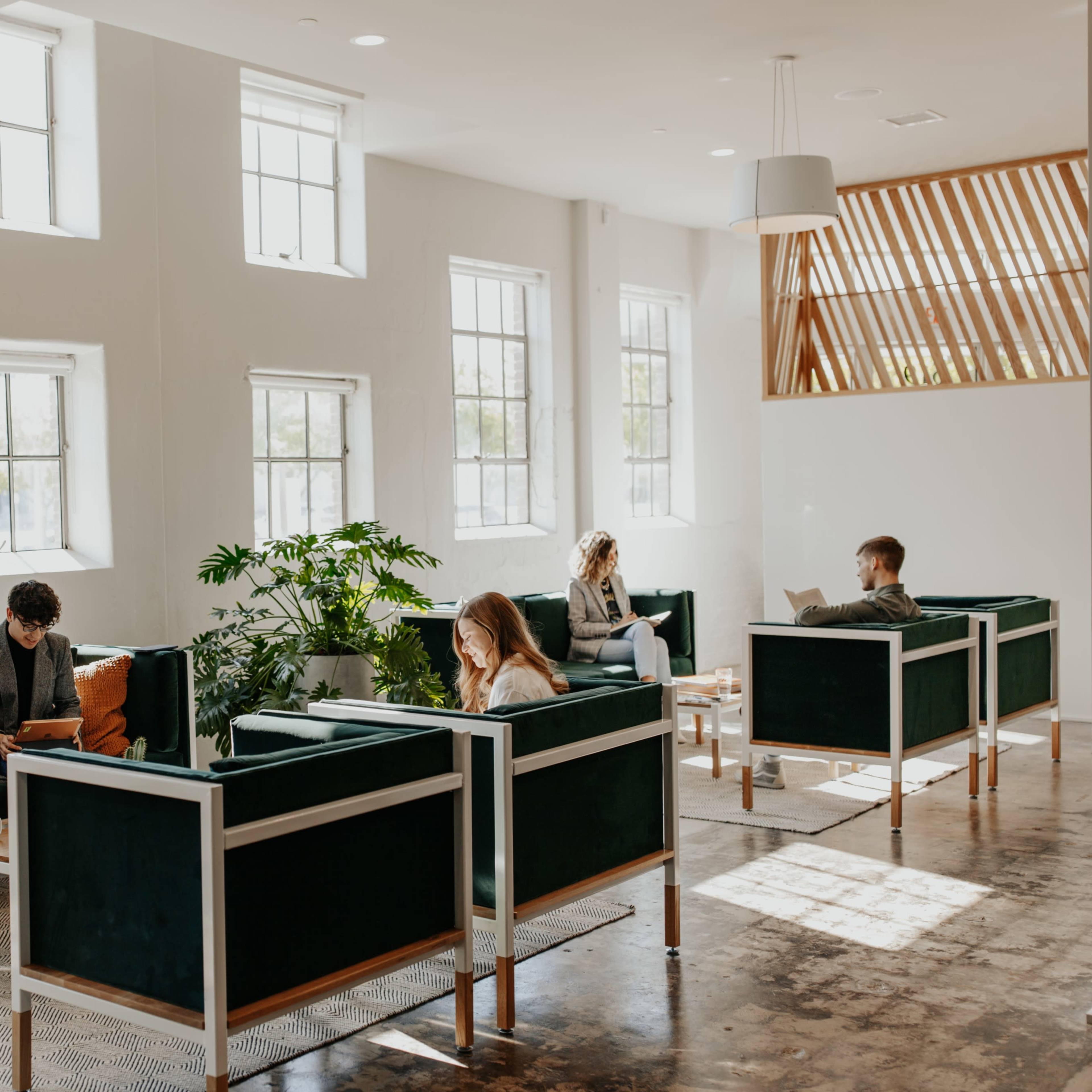 A group of individuals are seated in a modern, well-lit lounge area, engaging in reading and conversation around green upholstered sofas and large windows.