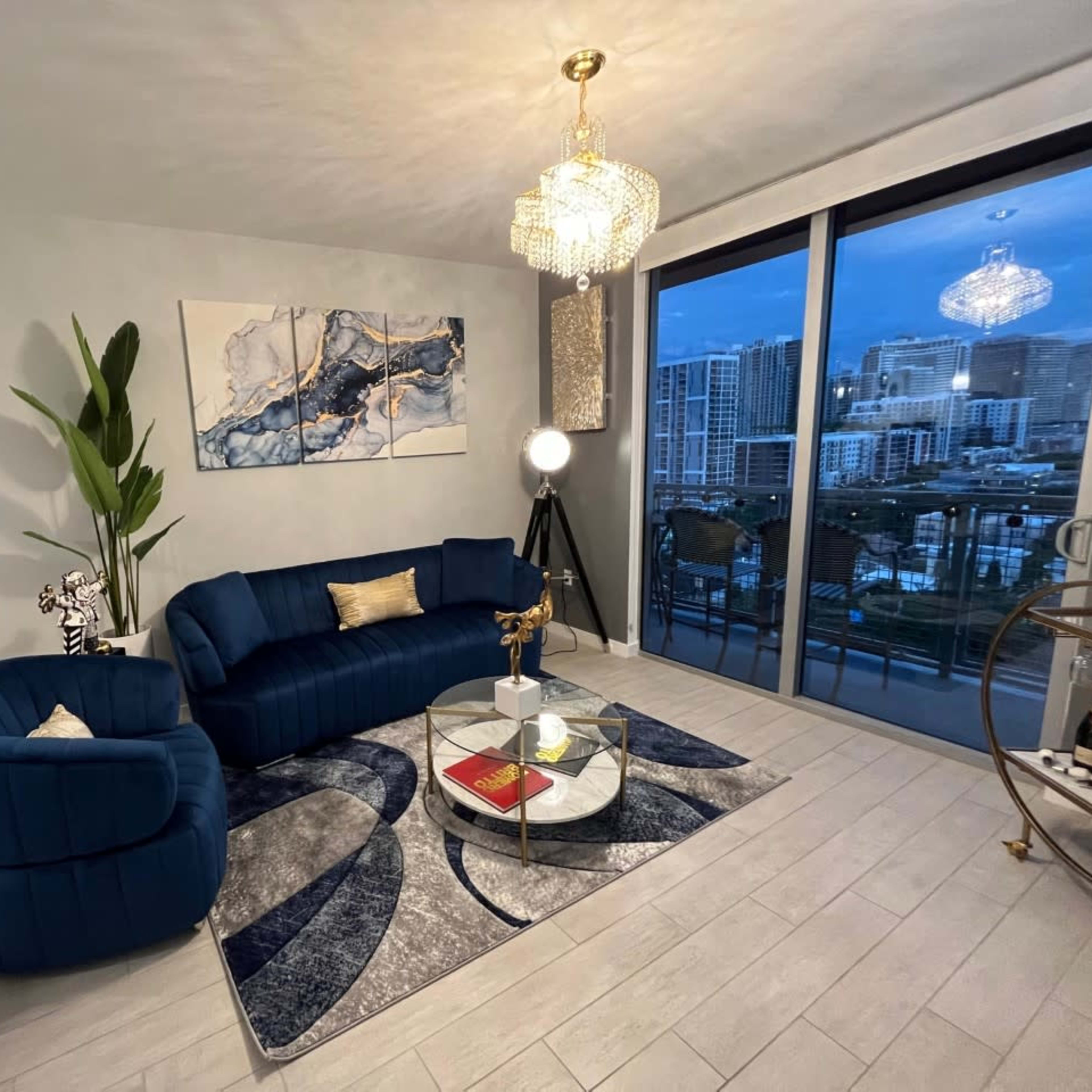 A stylish living room features a blue velvet sofa, a round glass coffee table, and large windows overlooking a cityscape.