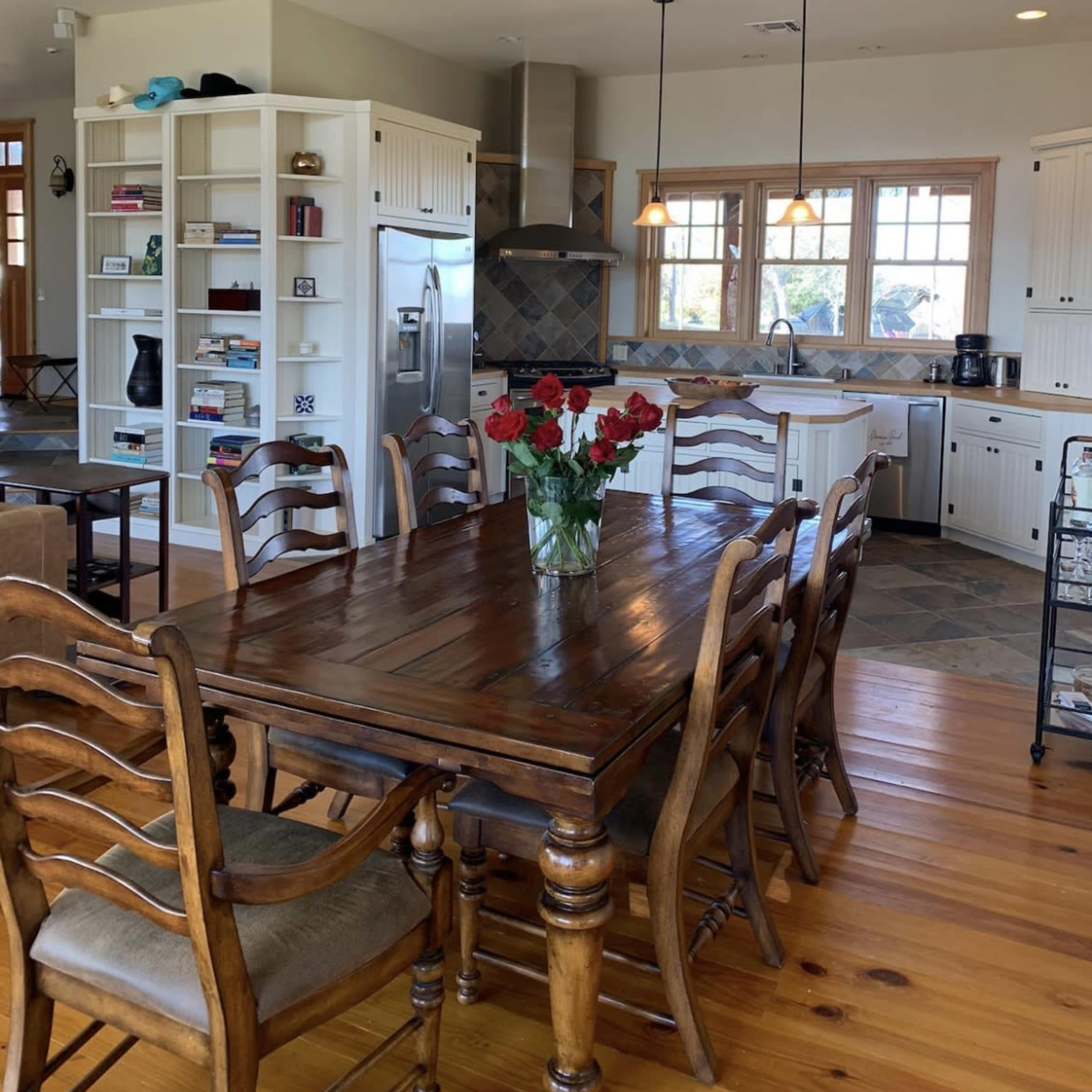 A spacious kitchen and dining area featuring a wooden dining table with chairs, a vase of red roses, white cabinets, and a stainless steel refrigerator.