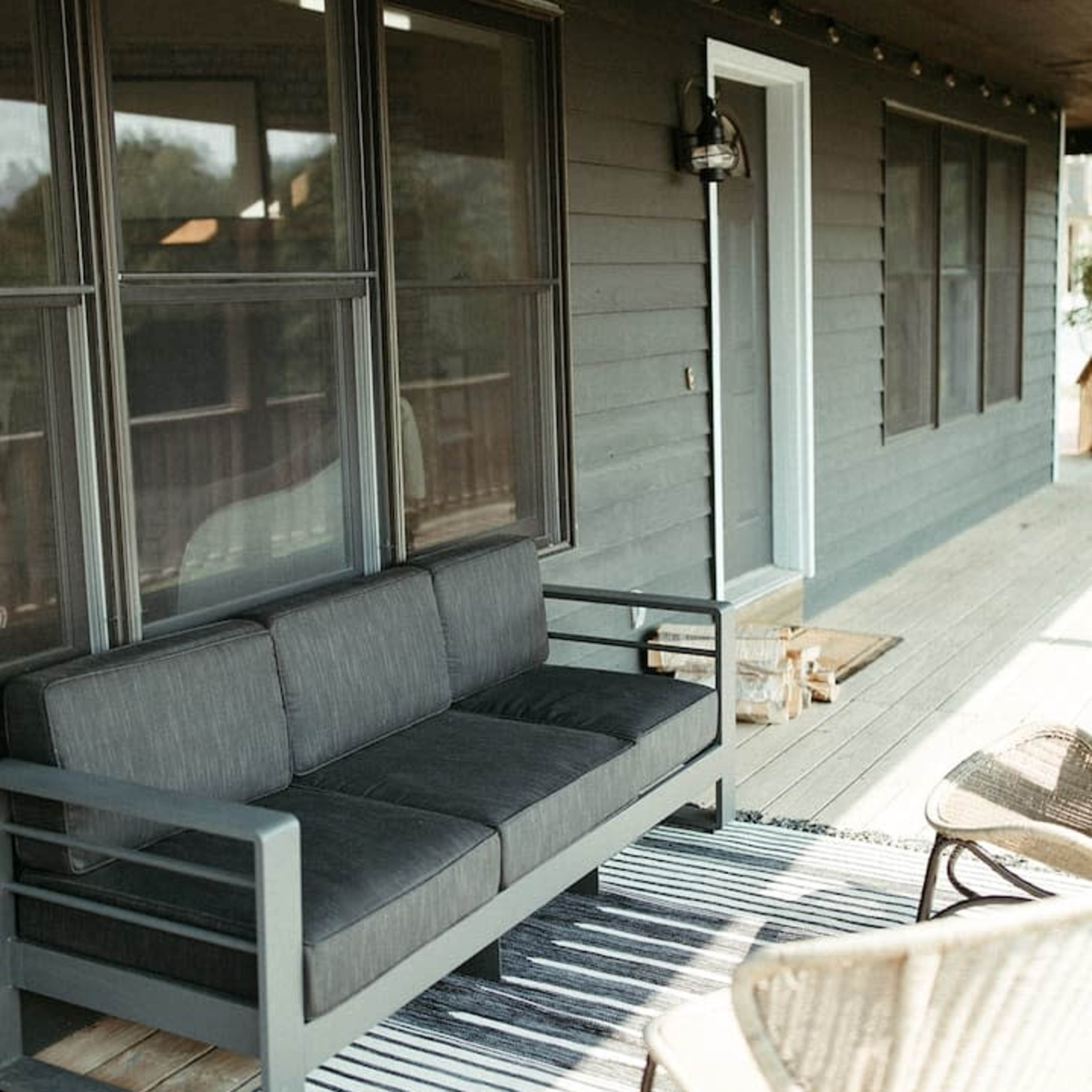 A gray outdoor sofa sits on a wooden porch beside a striped area rug and two wicker chairs.