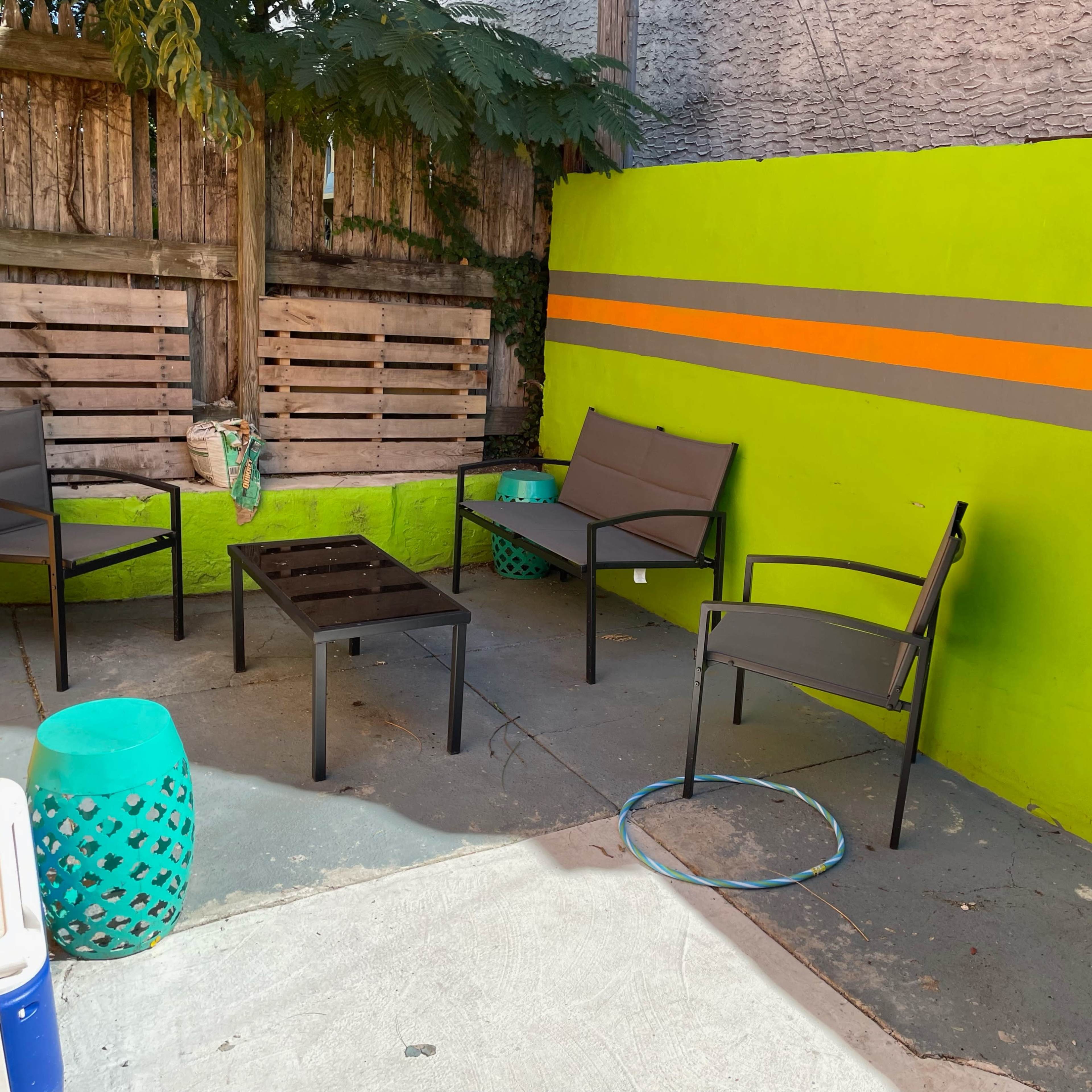 A small outdoor sitting area features four chairs arranged around a central table, with a colorful wall and potted plants in the background.