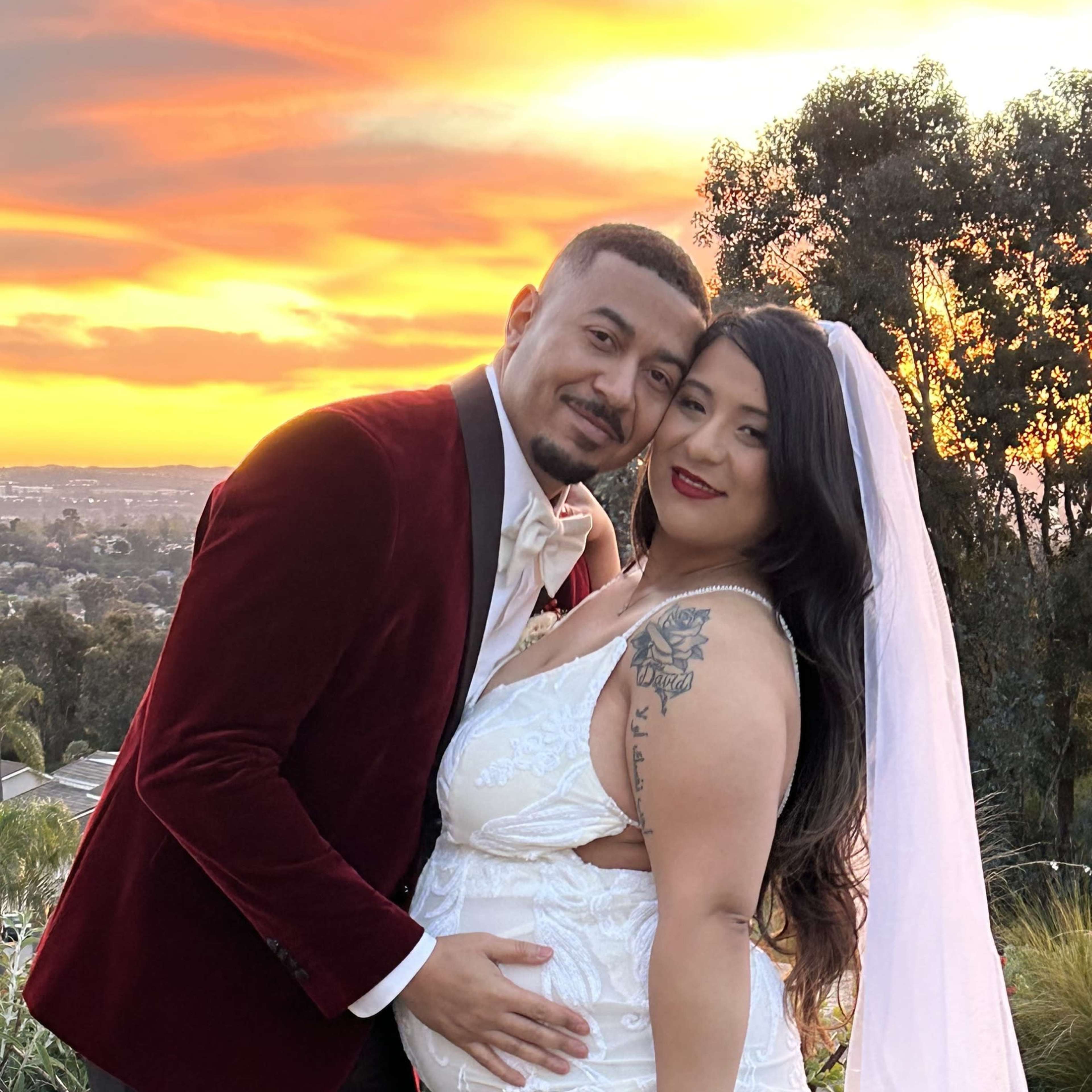 A couple poses together outdoors at sunset, with the woman wearing a white wedding gown and showing her baby bump, while the man is dressed in a red velvet suit.