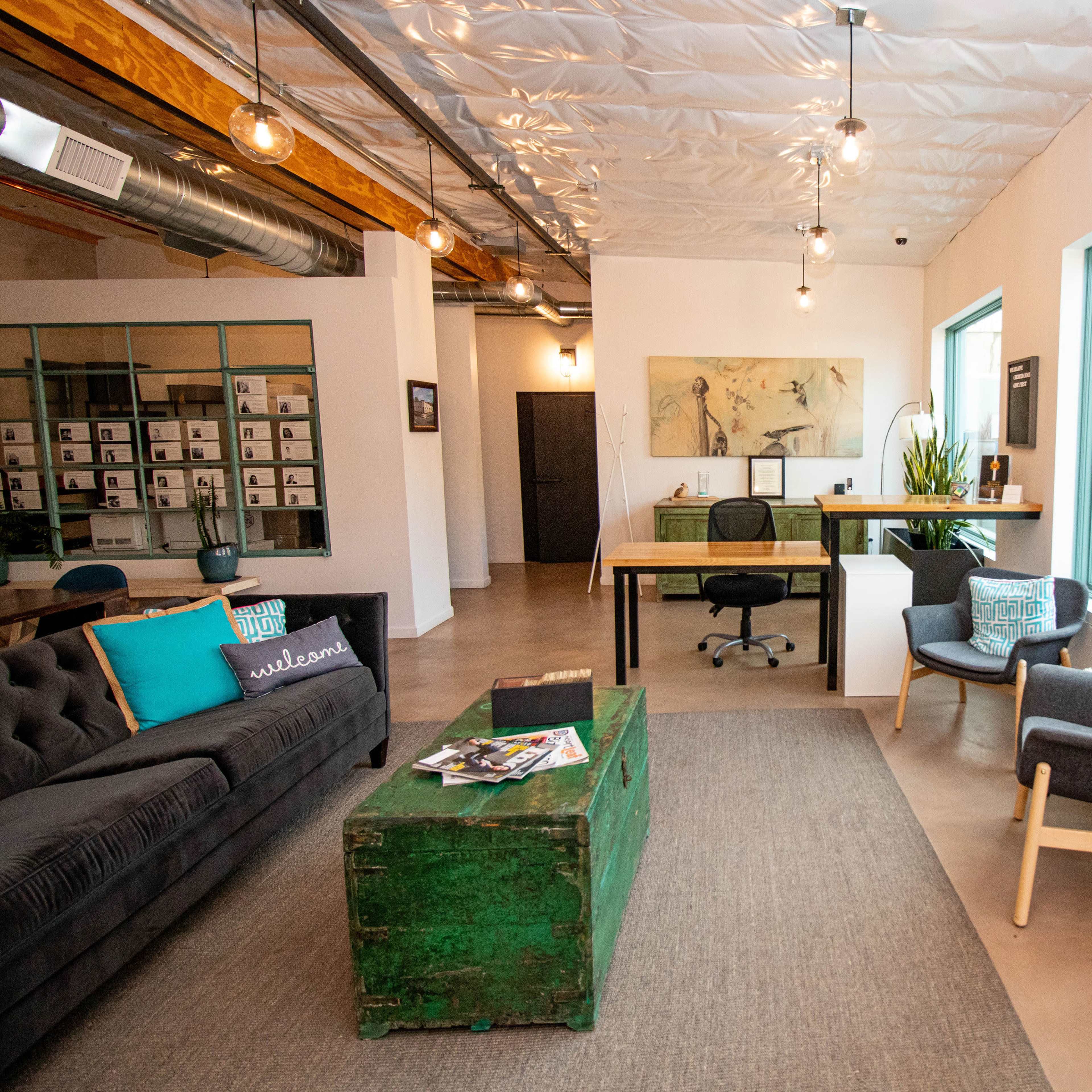 A modern office space featuring a green wooden coffee table, a gray sofa with decorative pillows, a desk with a chair, and large windows allowing natural light to enter.