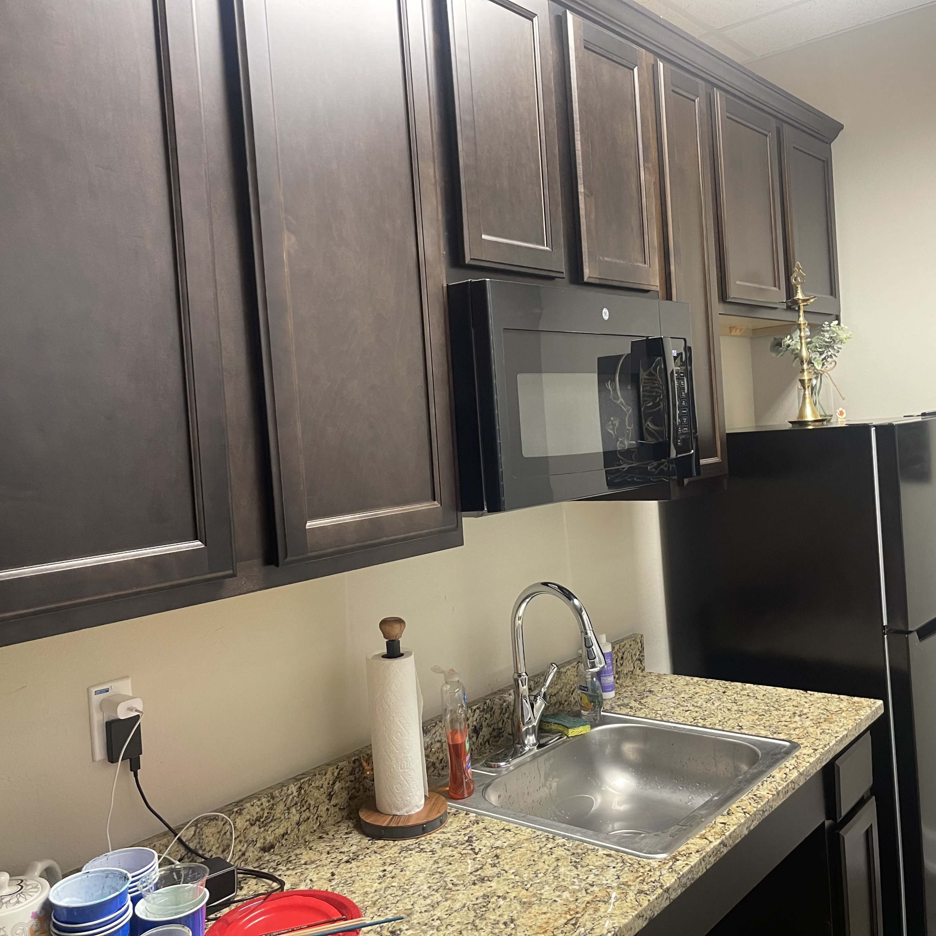 A kitchen with dark wooden cabinets, a stainless steel microwave, a black refrigerator, and a granite countertop featuring a sink and various dishes.