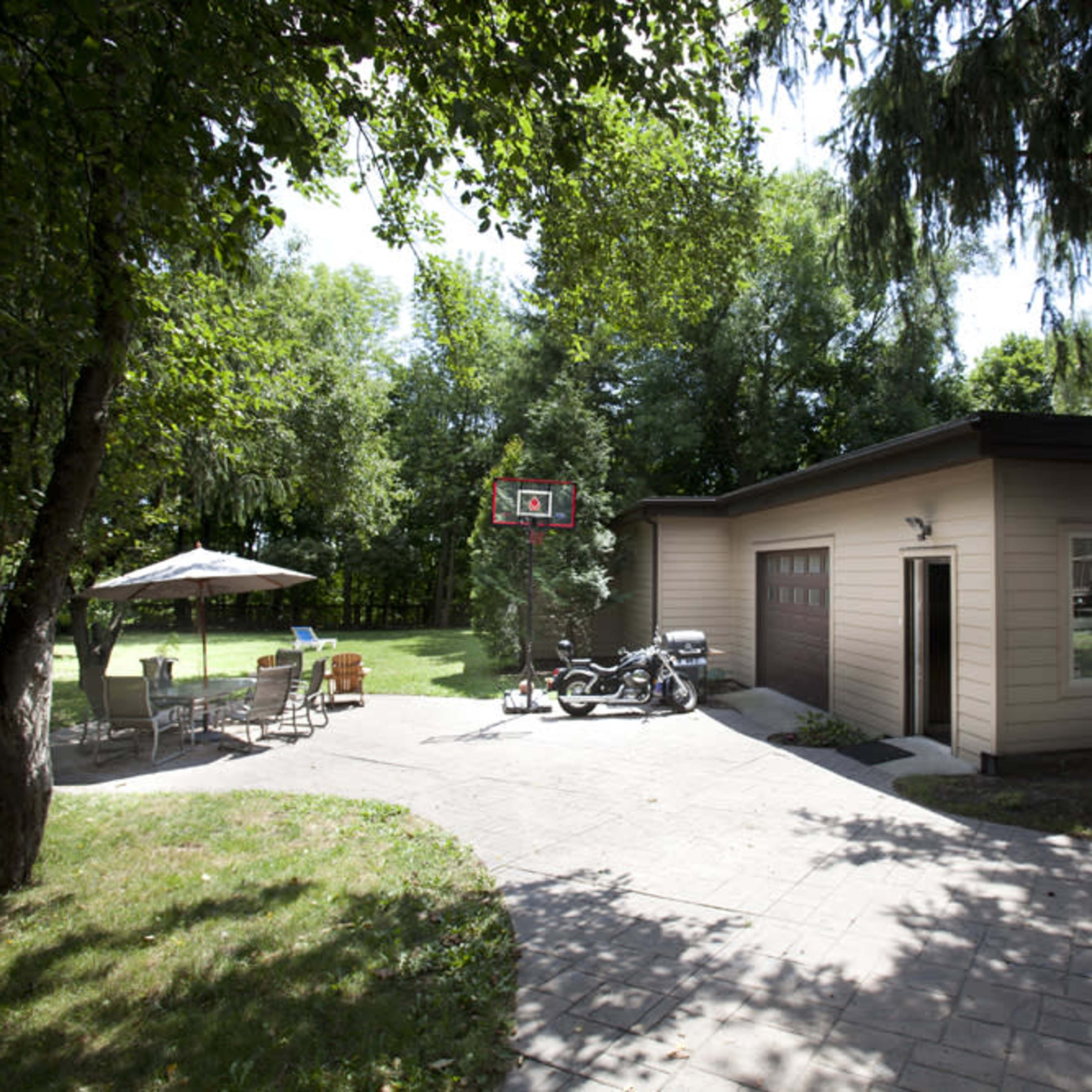 A residential backyard with a paved path leading to a garage, surrounded by trees, a patio set, and a basketball hoop.