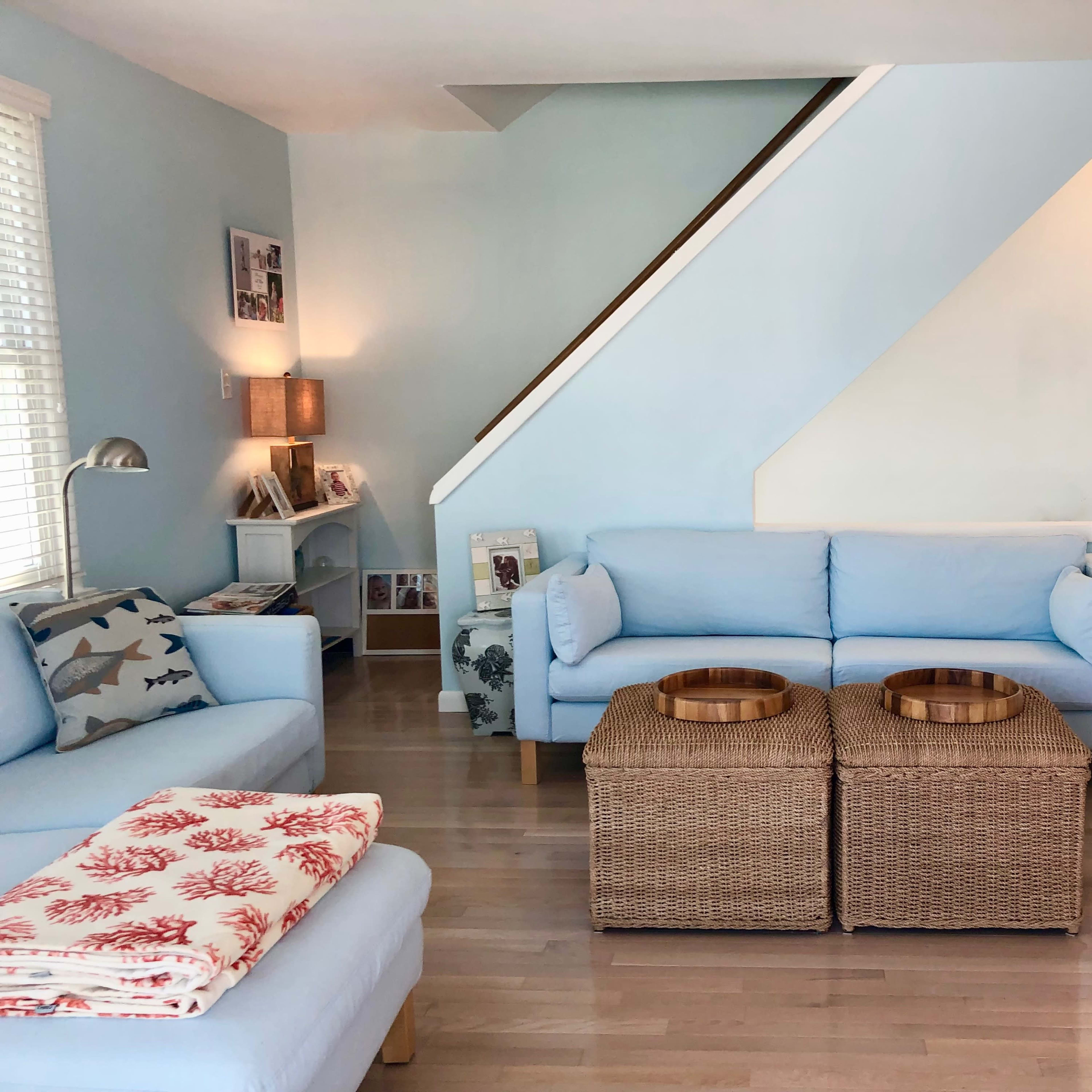 A living room features light blue walls, a staircase, two wicker coffee tables, and soft sofas arranged around a cozy seating area.