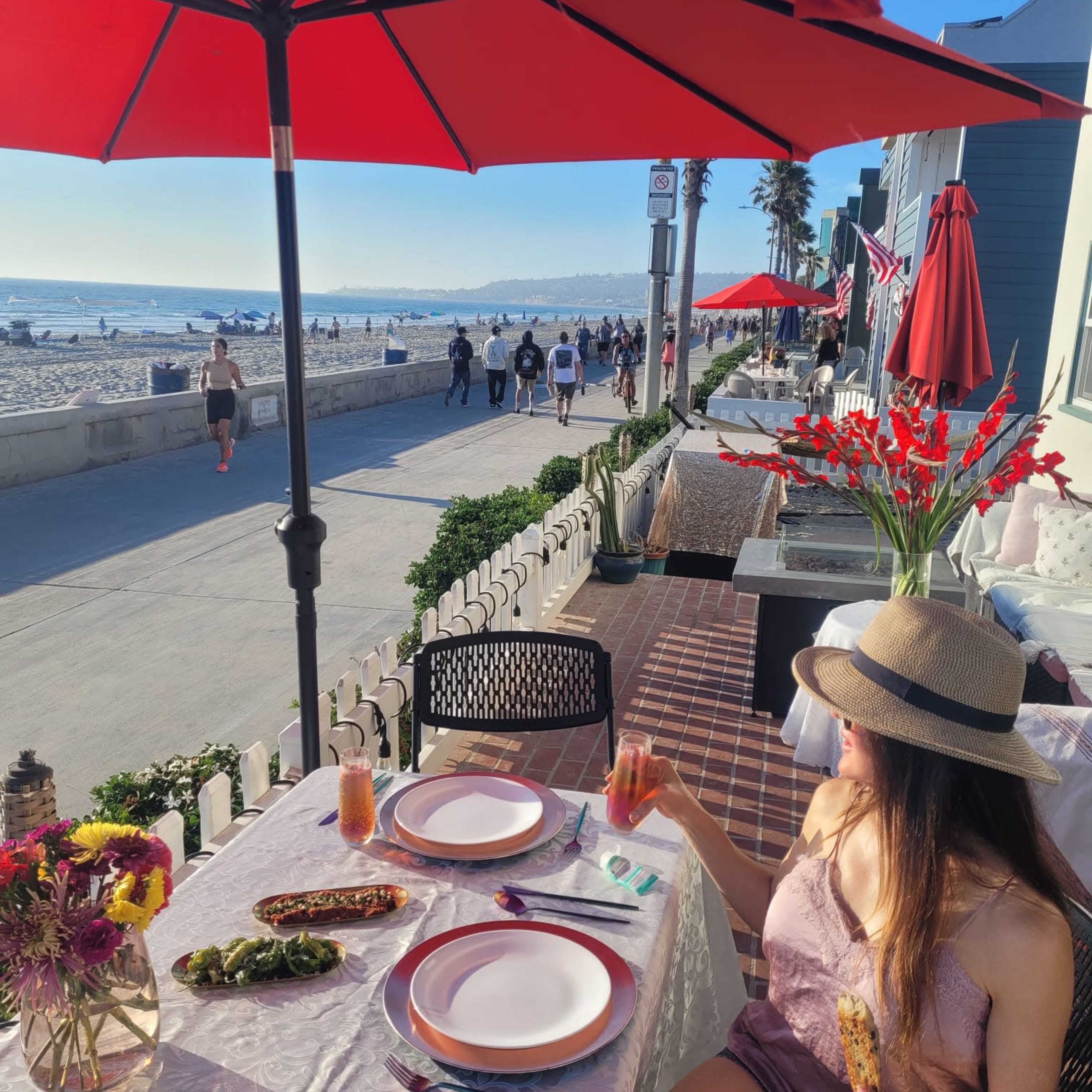A woman sits at a table set for a meal on a patio overlooking a beach promenade, with flowers and drinks on the table and umbrellas providing shade.