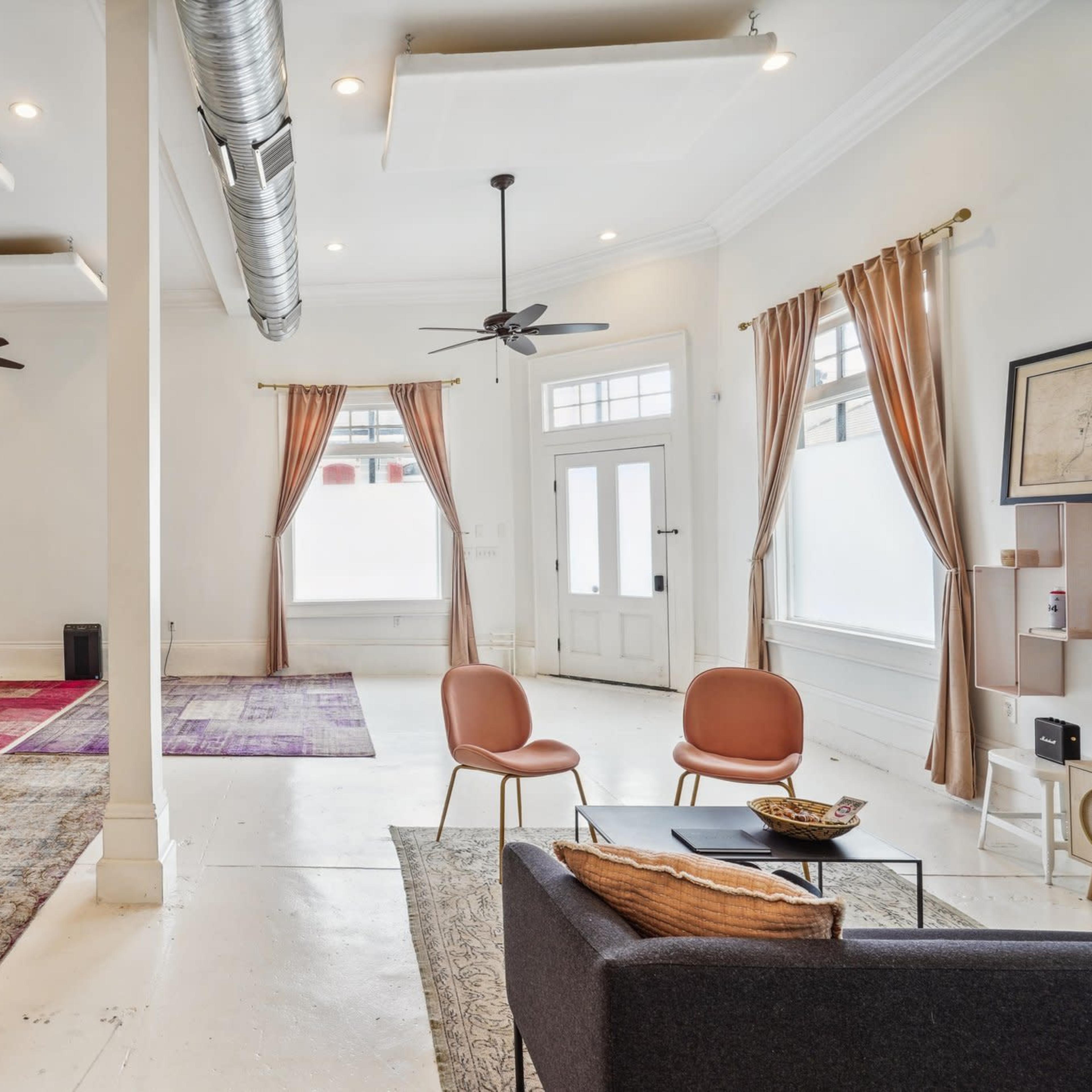 A spacious, minimally furnished room with large windows, two ceiling fans, and a mix of rugs on the polished floor.