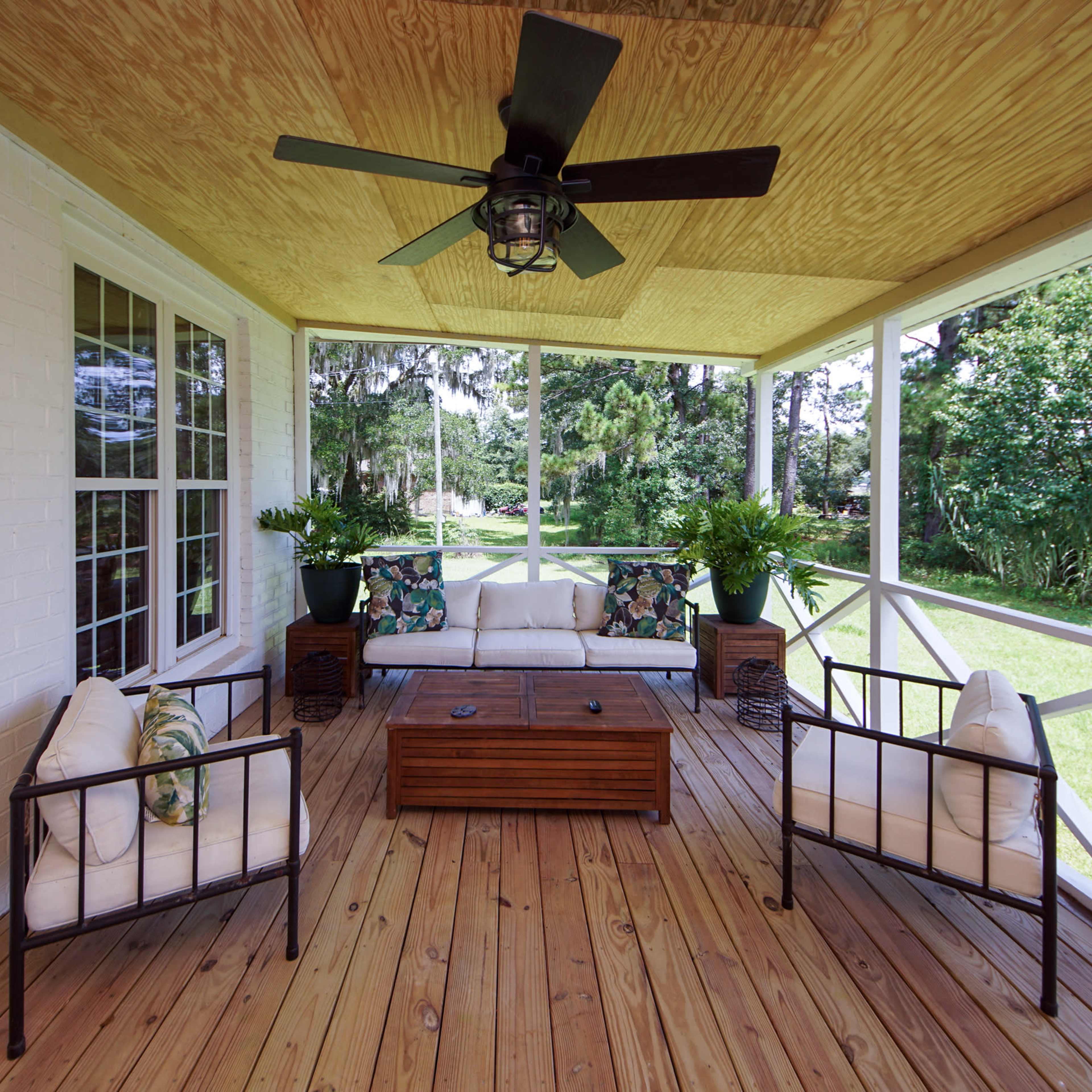A covered porch with a wooden floor, two metal-framed chairs, a coffee table, and a view of greenery outside.