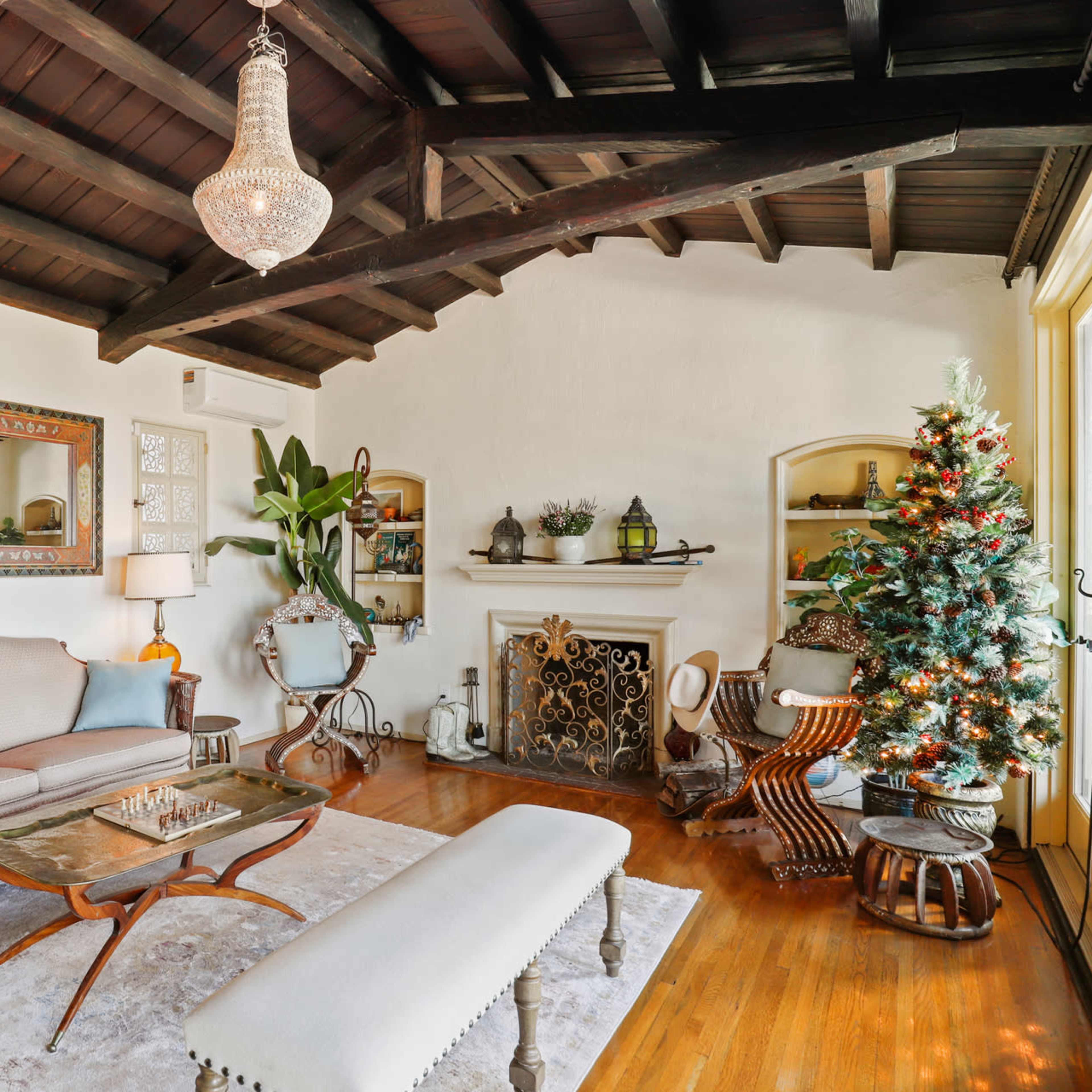 A cozy living room features a beige sofa, a glass coffee table, and a decorated Christmas tree beside a fireplace, complemented by wooden beams on the ceiling.