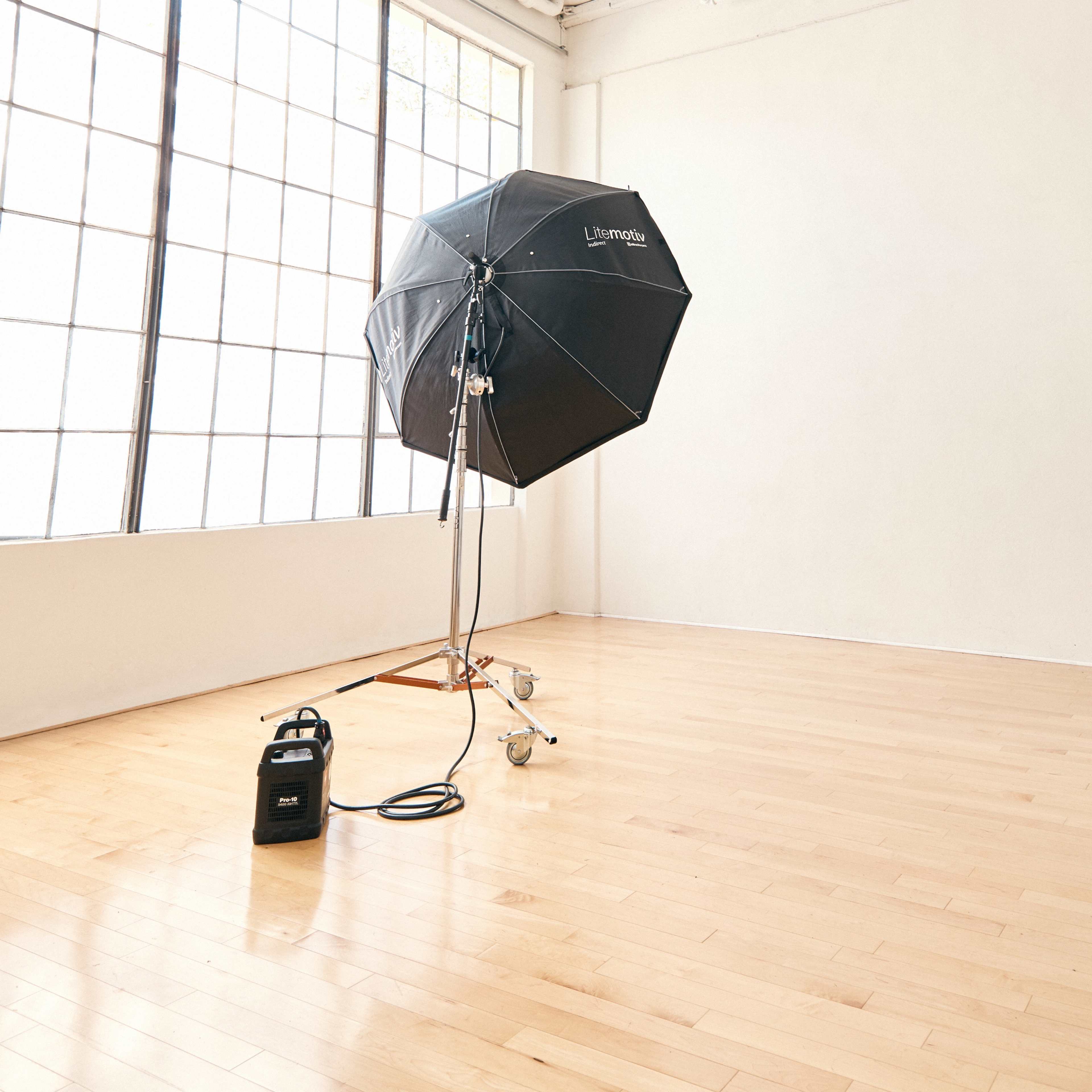 A black photography umbrella light stands on a wheeled stand in an empty, well-lit studio with wooden floors and large windows.