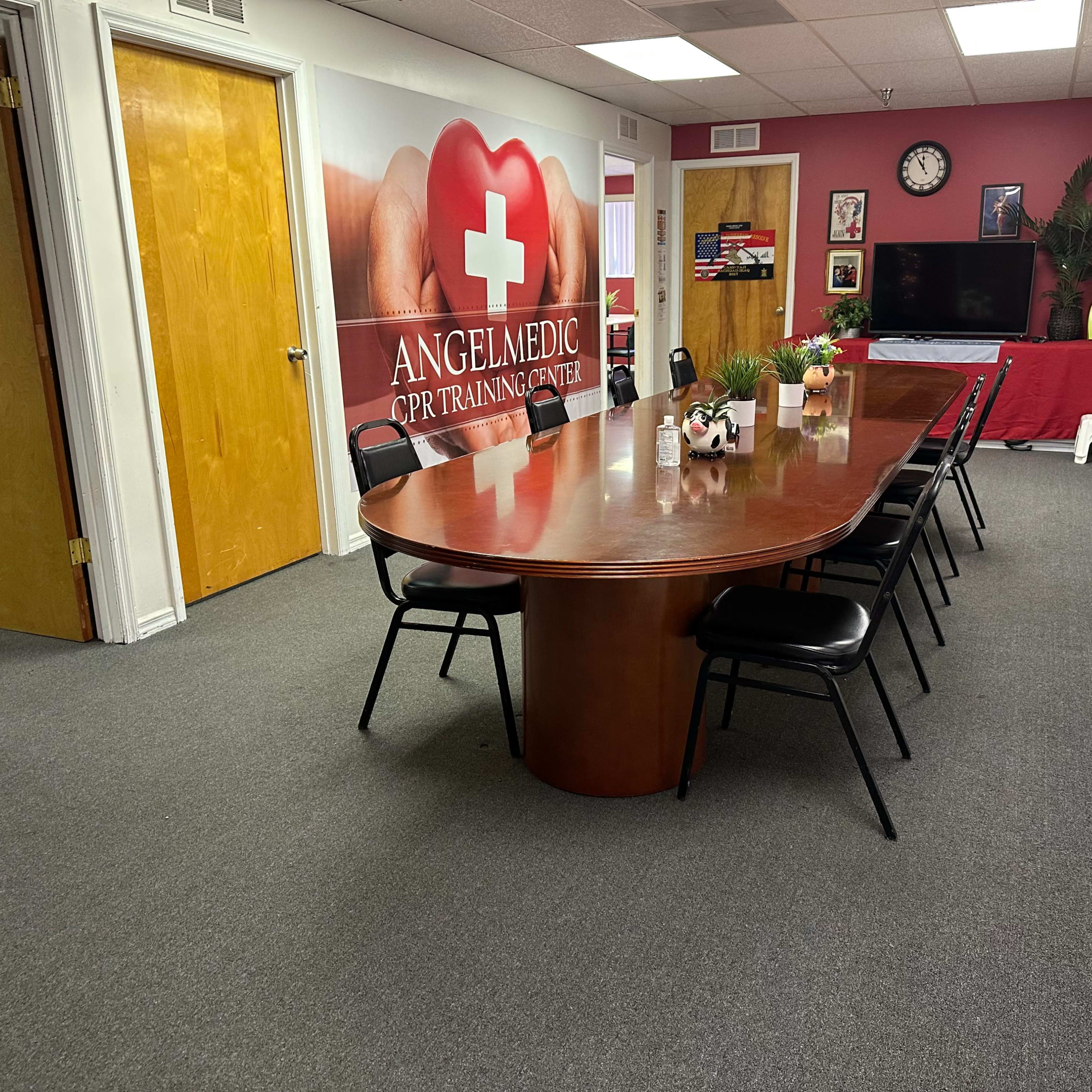 A training room features a long wooden conference table surrounded by black chairs, with a wall displaying a large red cross and medical images.