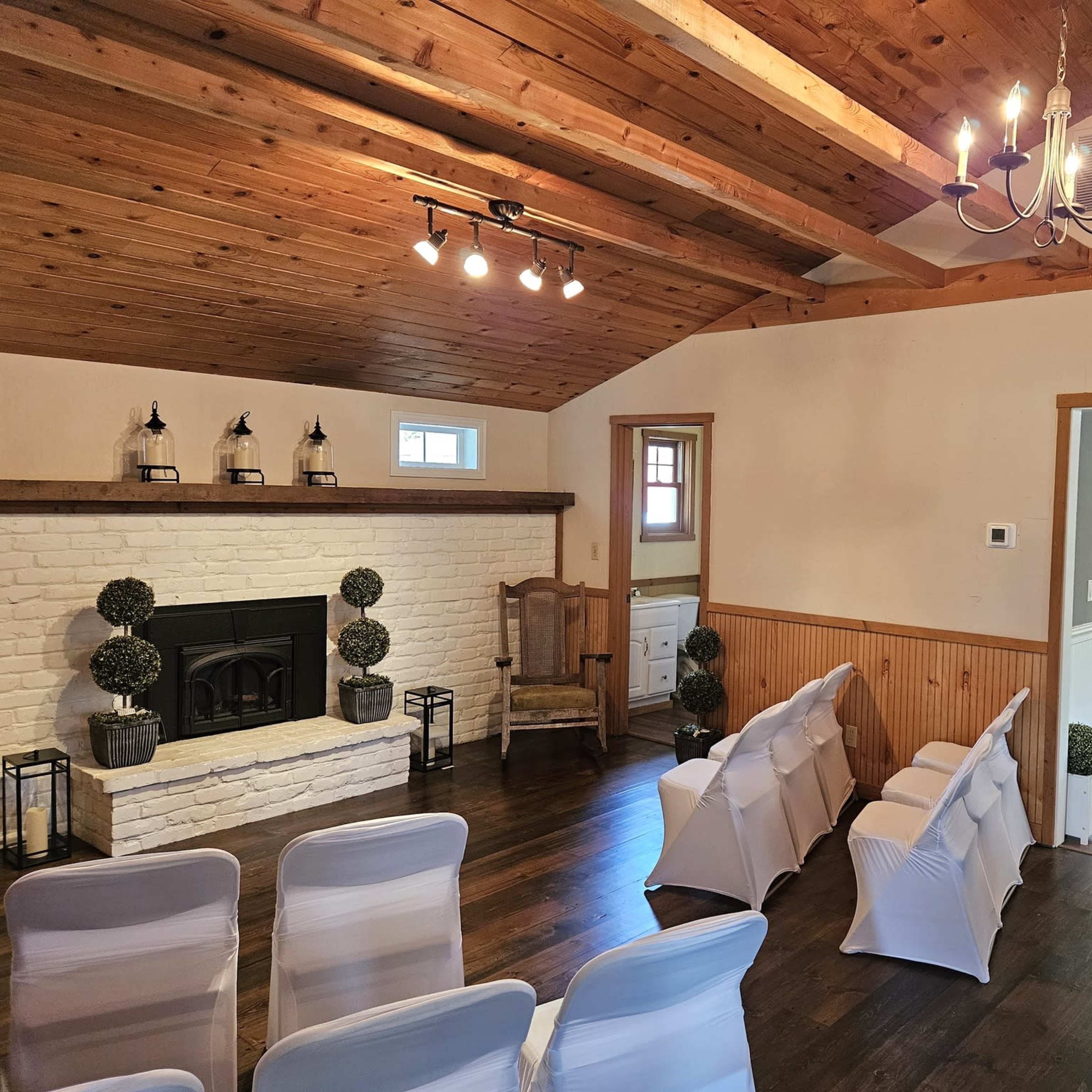 An indoor ceremony space featuring white chairs arranged in front of a brick fireplace, with wooden beams on the ceiling and a table visible in the adjacent room.