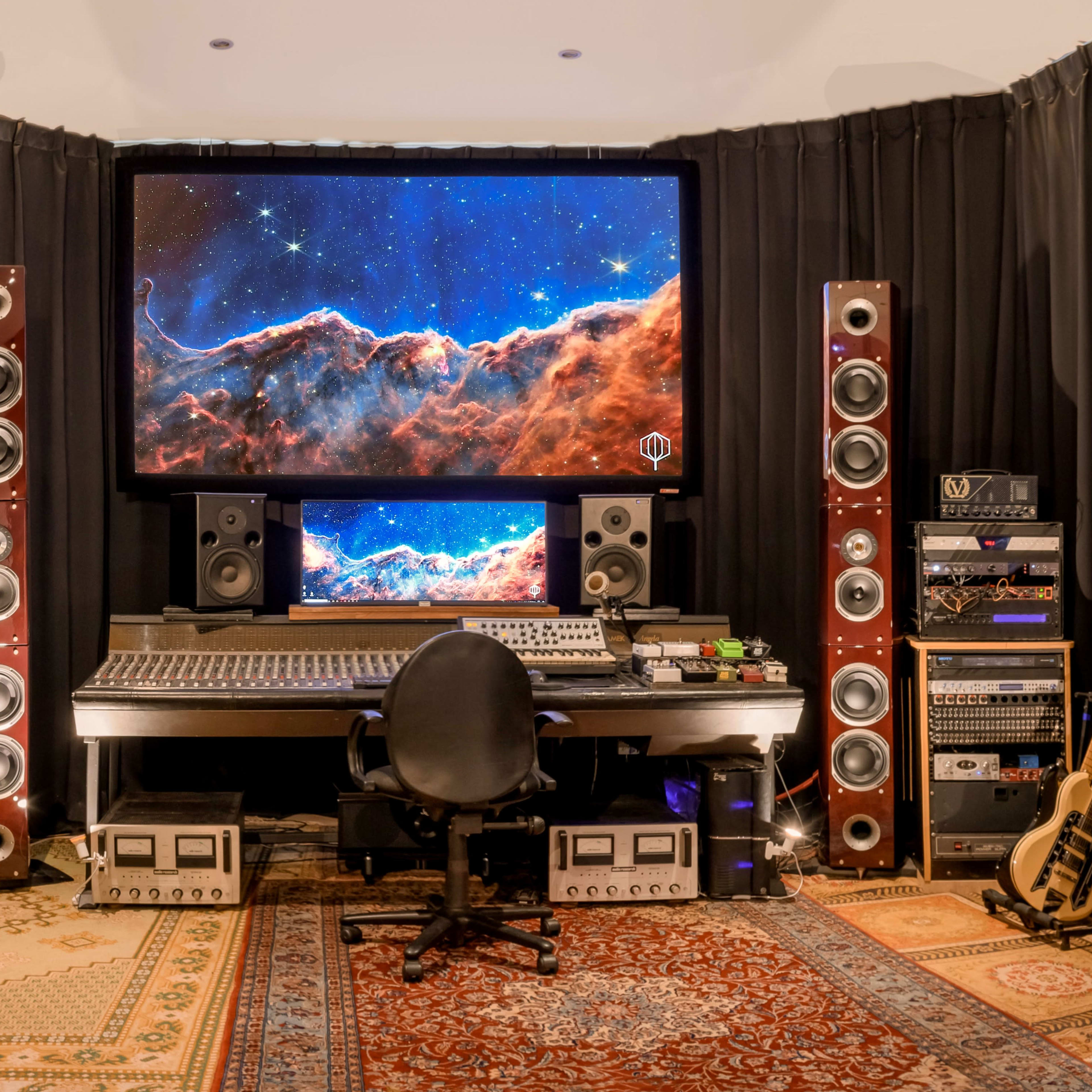 A modern music production studio features a large central workstation with two screens displaying a cosmic scene, surrounded by tall speakers and various music equipment.