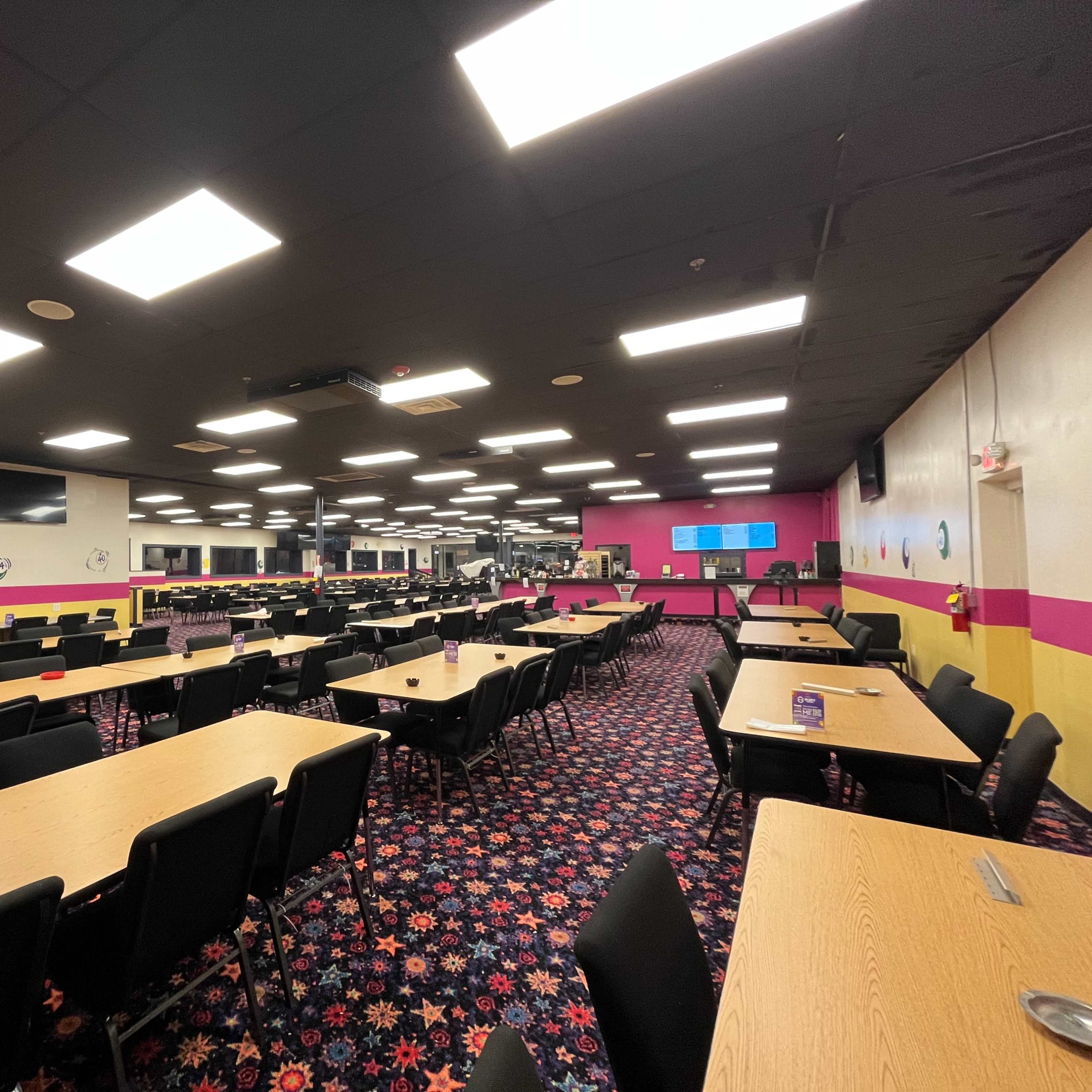 A spacious dining area with many wooden tables and black chairs arranged in rows, featuring a colorful carpet and bright overhead lighting.