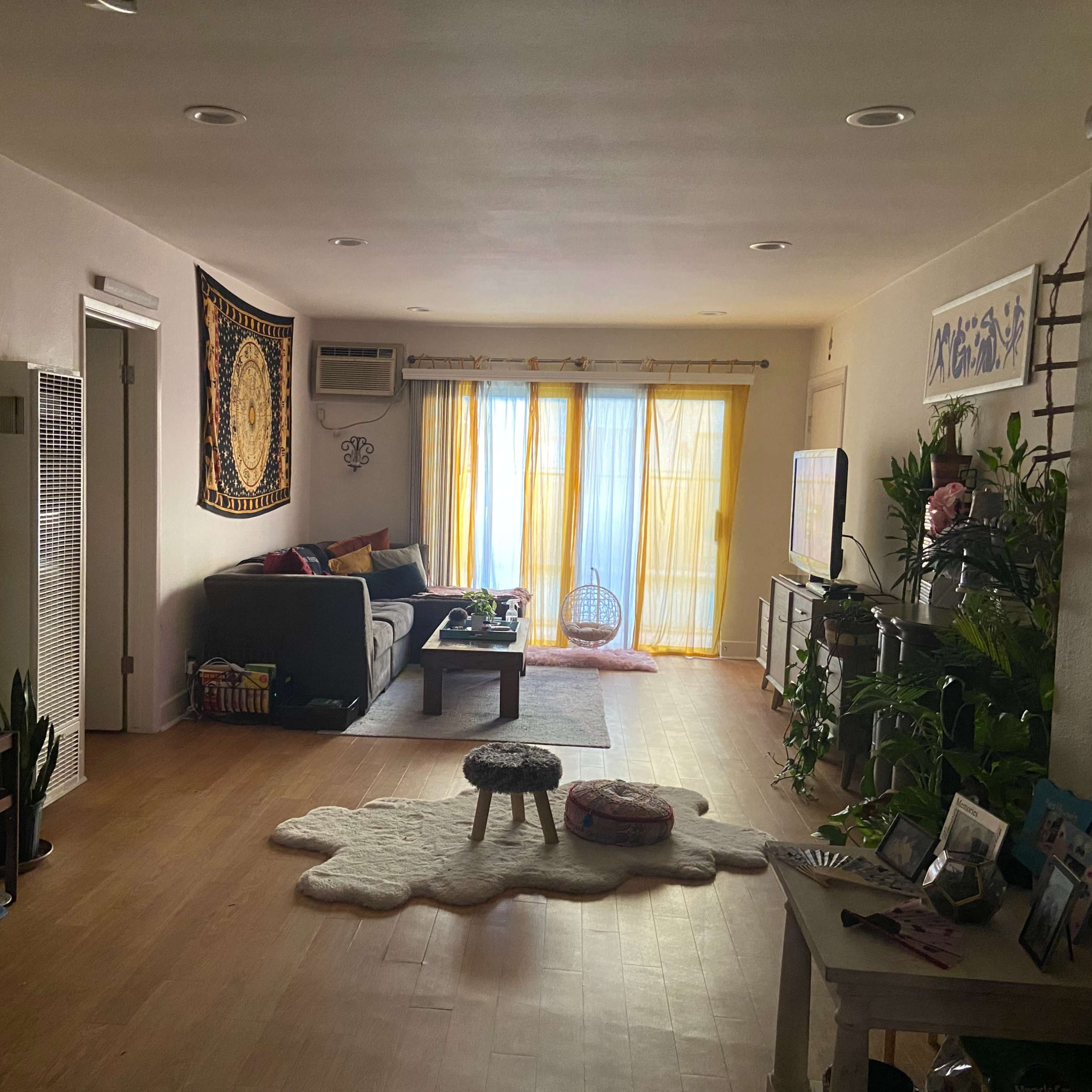 A spacious living room features a couch, plants, and large windows with yellow curtains.