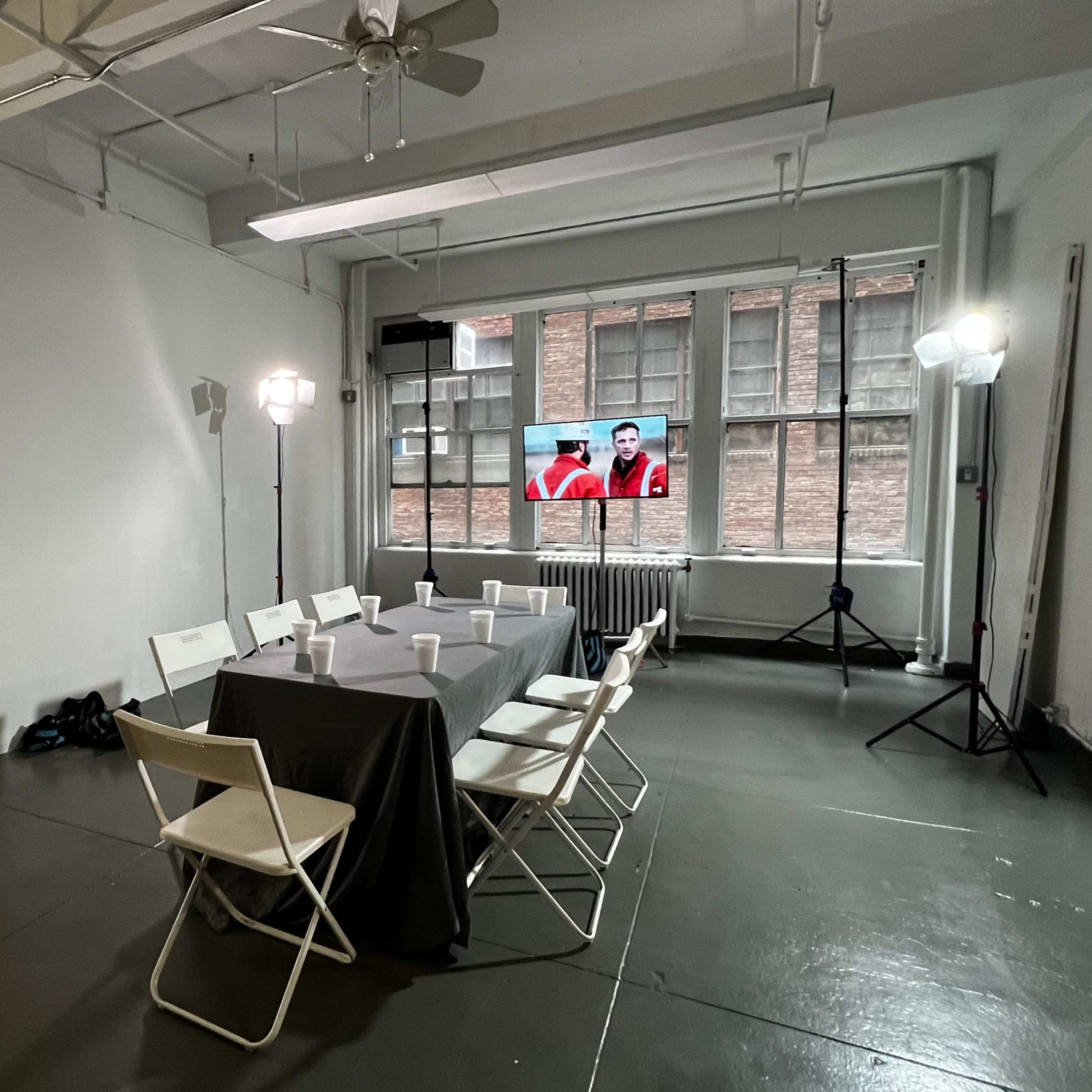 A well-lit meeting room with a table set for a presentation, featuring chairs, a television displaying a video, and a mix of lighting equipment.