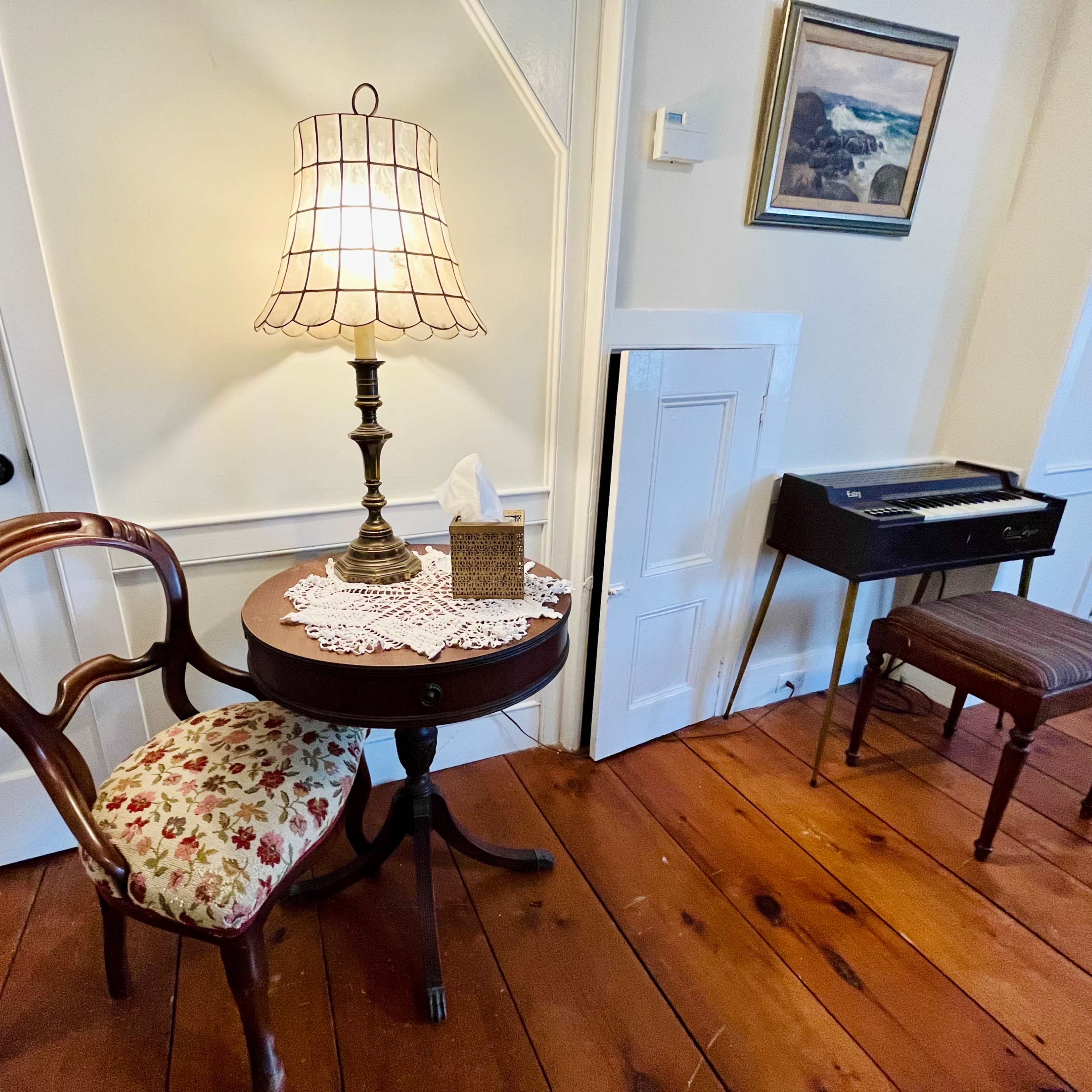 A small wooden table with a decorative lamp, a tissue box, and a chair is positioned beside a wall, with a musical keyboard and a stool visible in the background.