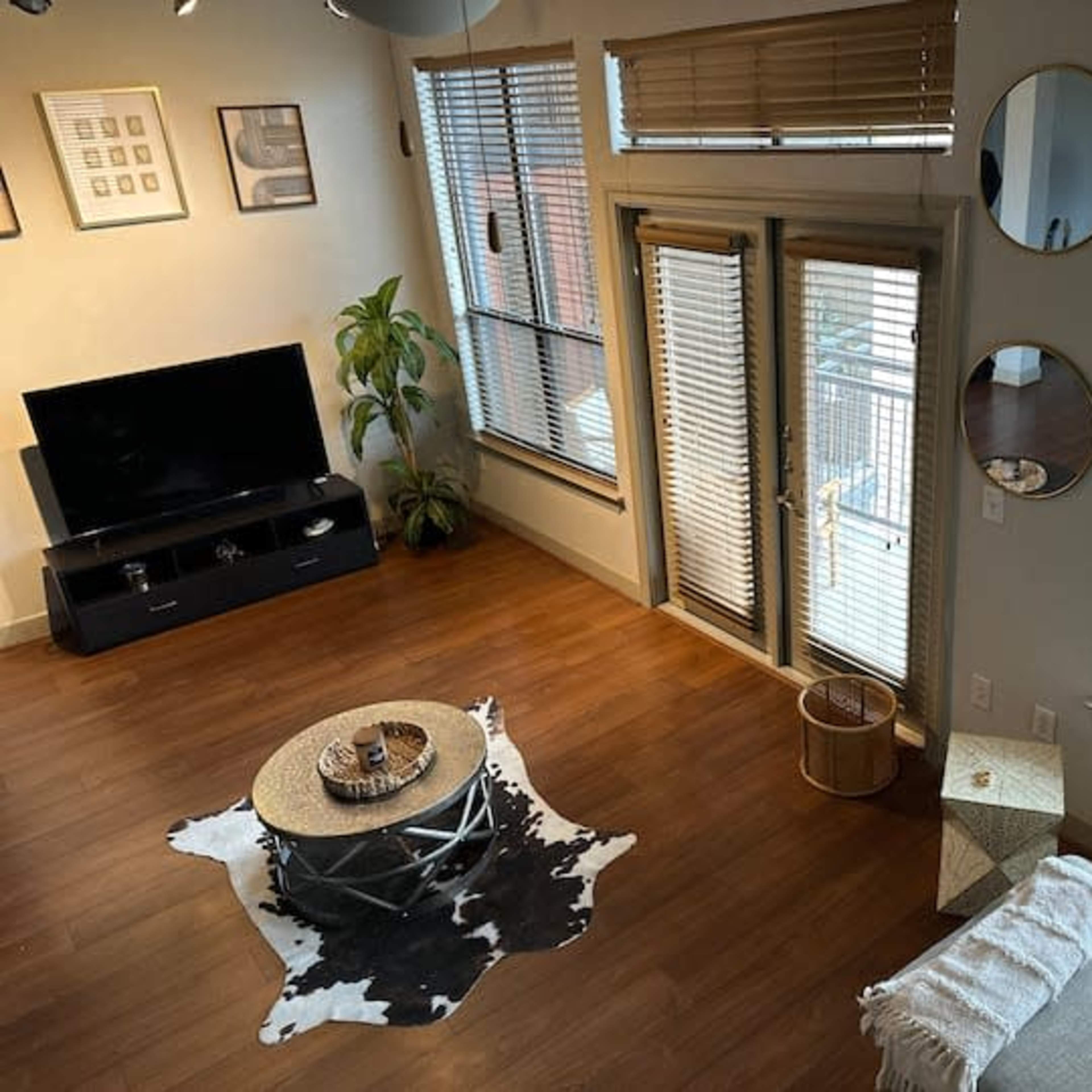 A living room features a television on a cabinet, a circular coffee table on a cowhide rug, a couch draped with a throw, and large windows with blinds.