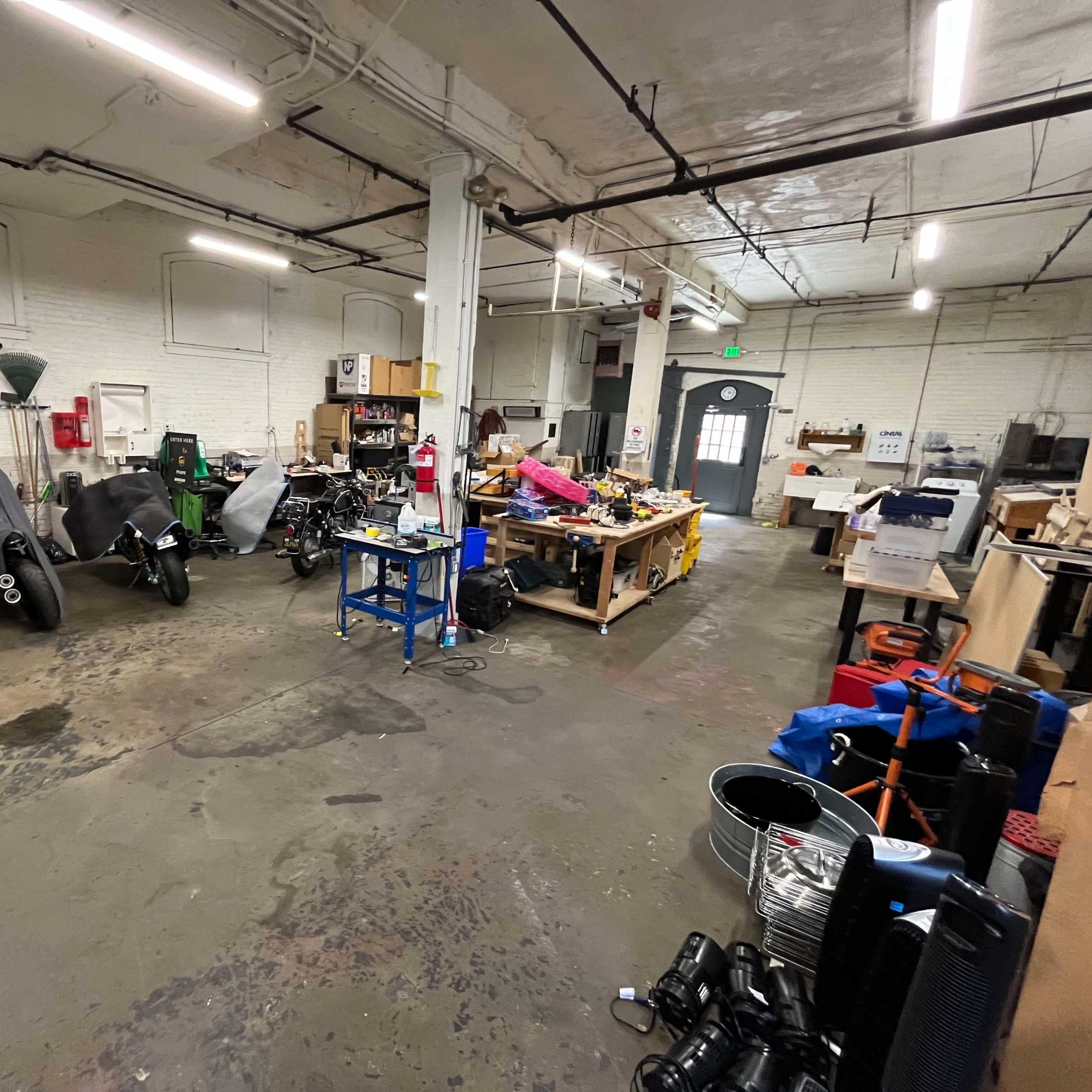 A spacious workshop contains motorcycles under covers, various tools, and cluttered work tables amidst an industrial setting.