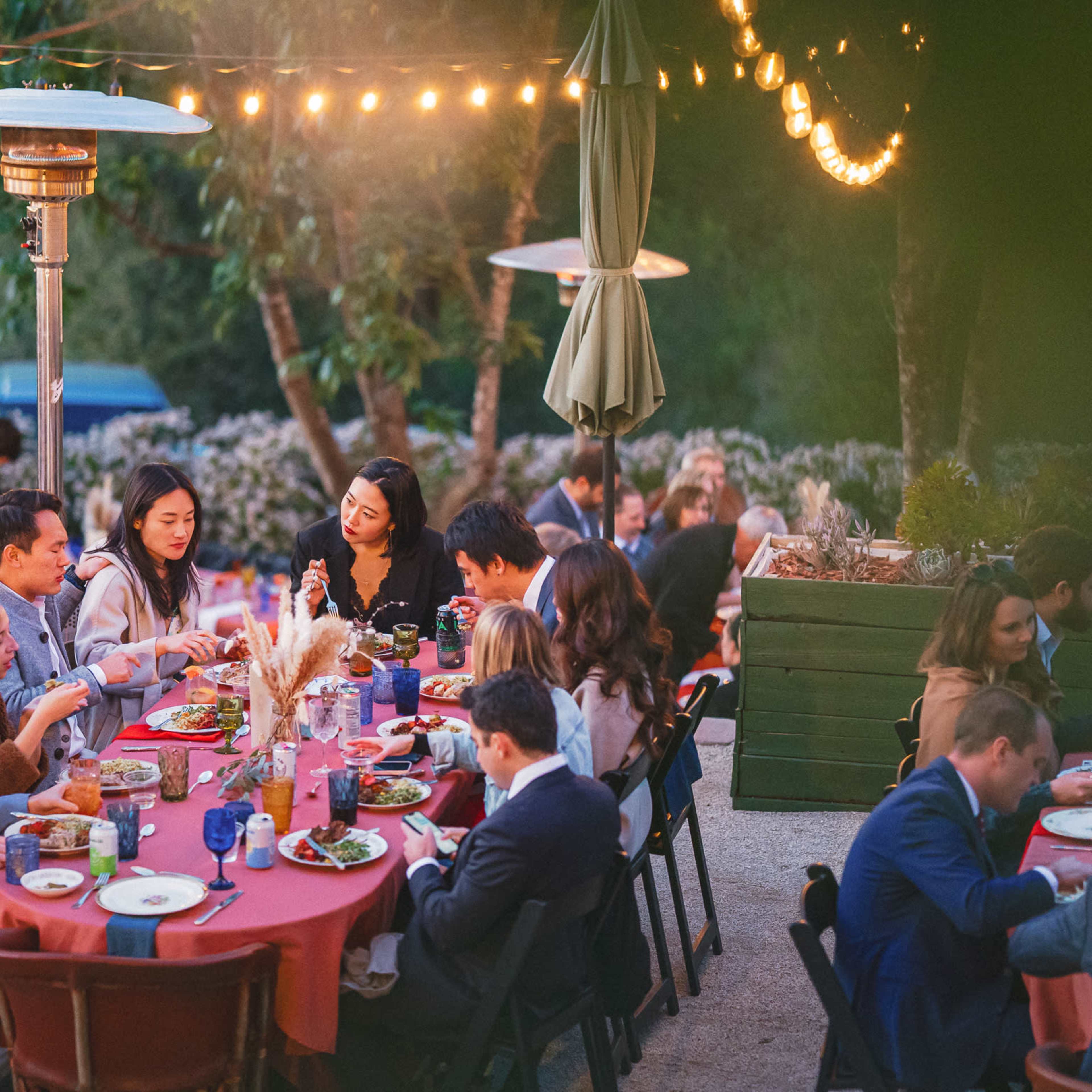 A group of people gather around outdoor tables enjoying a meal illuminated by string lights in a garden setting.