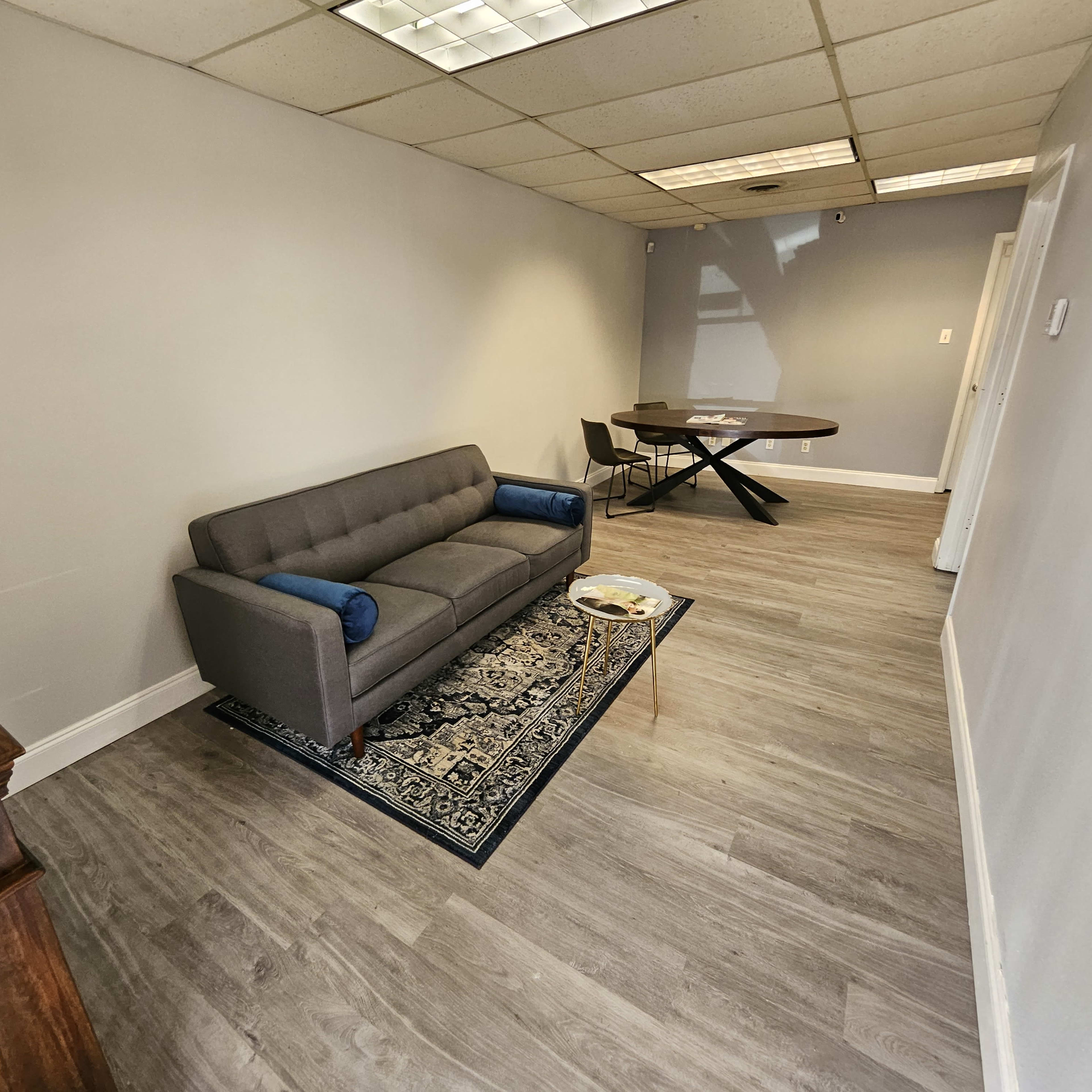 A sparsely furnished office space with a gray sofa, a round table, and a small decorative rug on a wooden floor.
