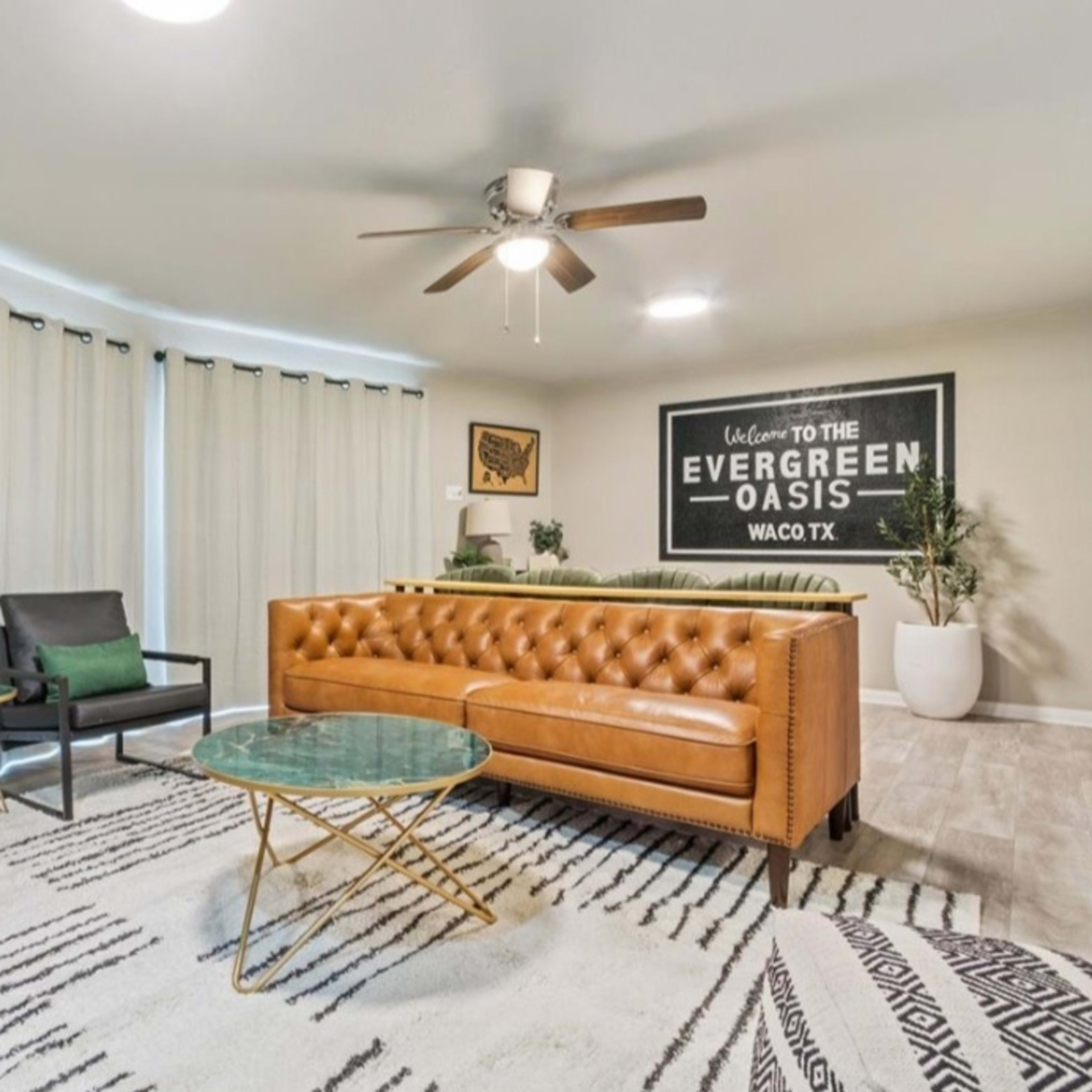 A living room with a tan tufted sofa, two black chairs, a glass coffee table, and a large wall sign that reads "Welcome to the Evergreen Oasis."