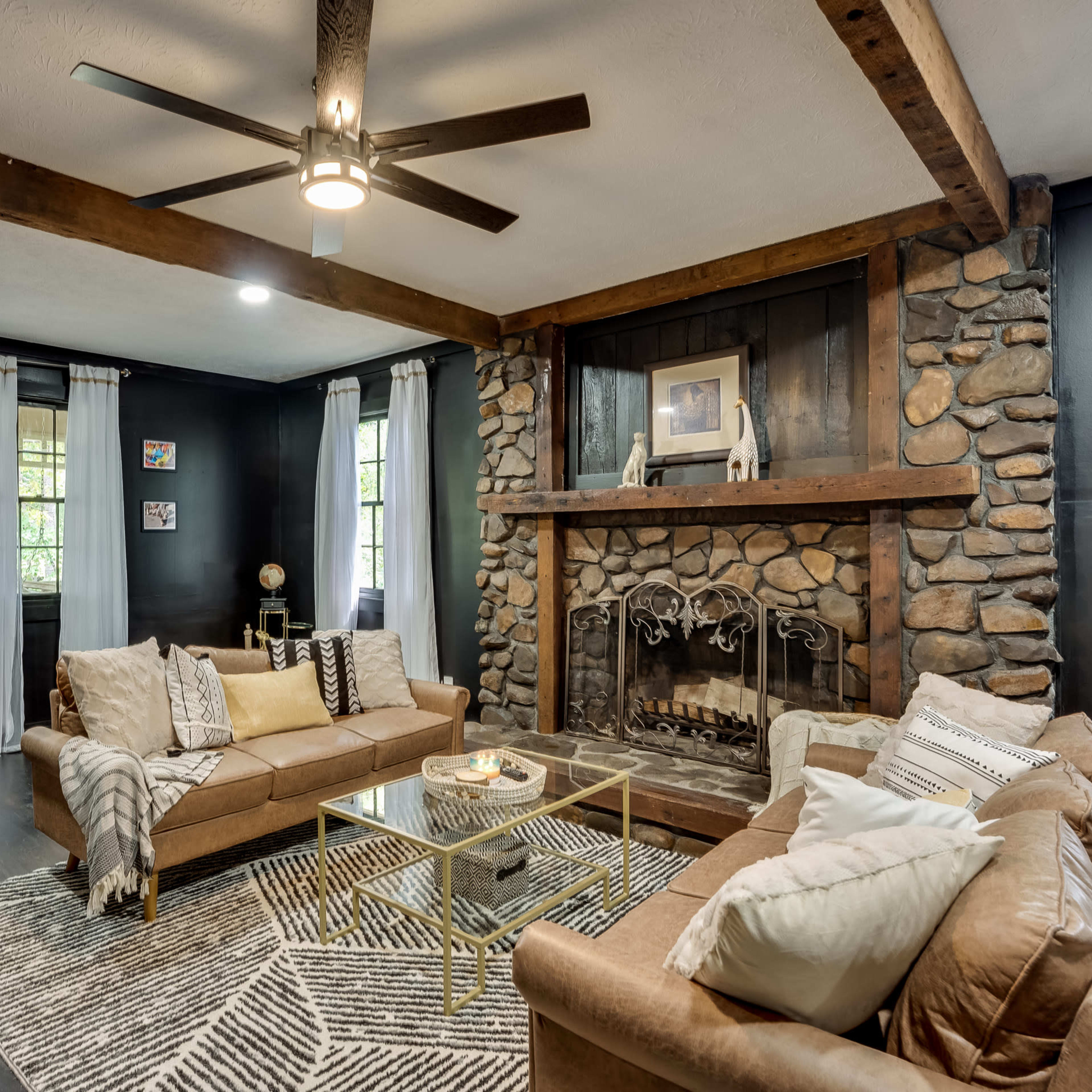 A living room with a stone fireplace, two brown couches, a glass coffee table, and large windows adorned with white curtains.