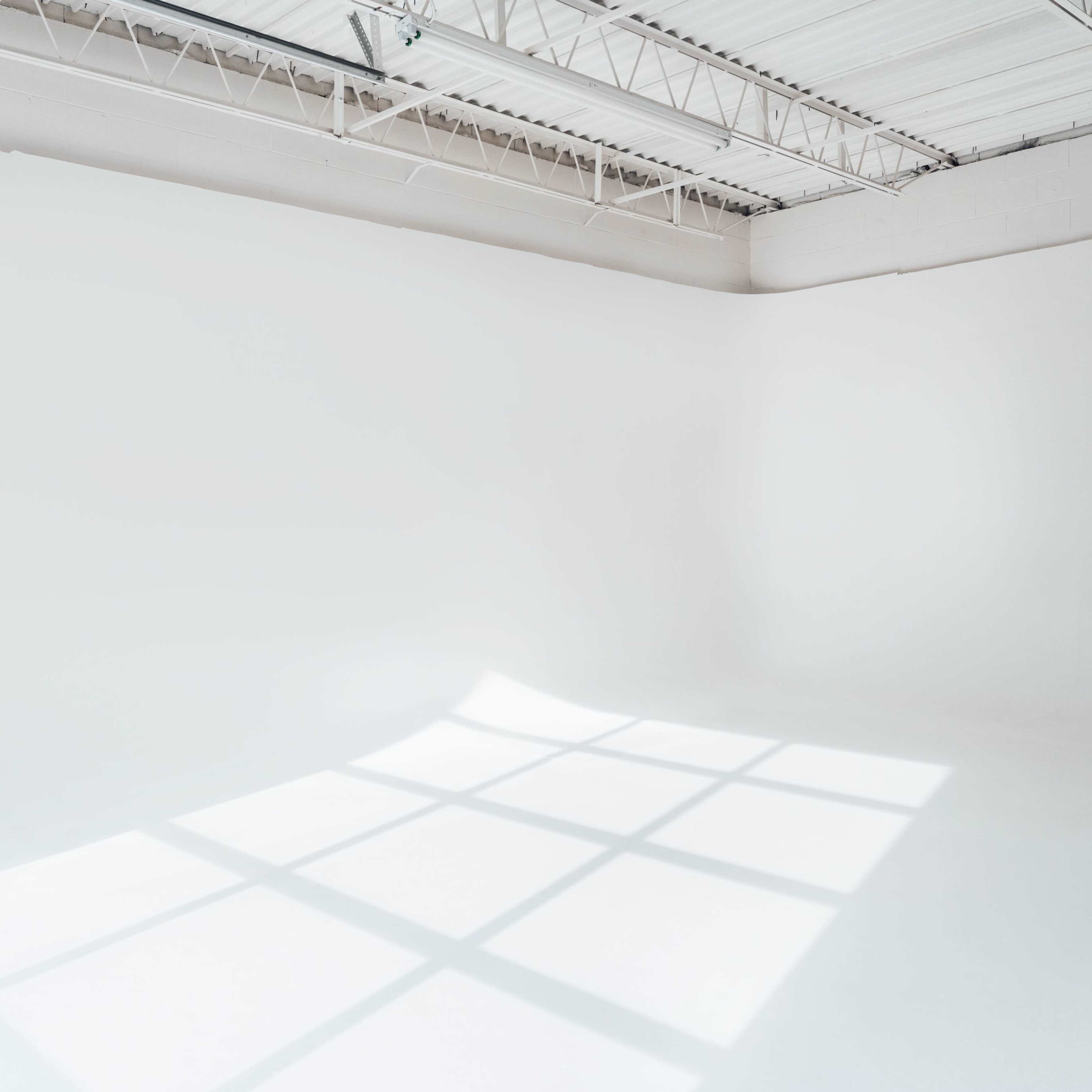 A large, empty studio space with white walls and a bright, illuminated floor creating shadows from window grids above.