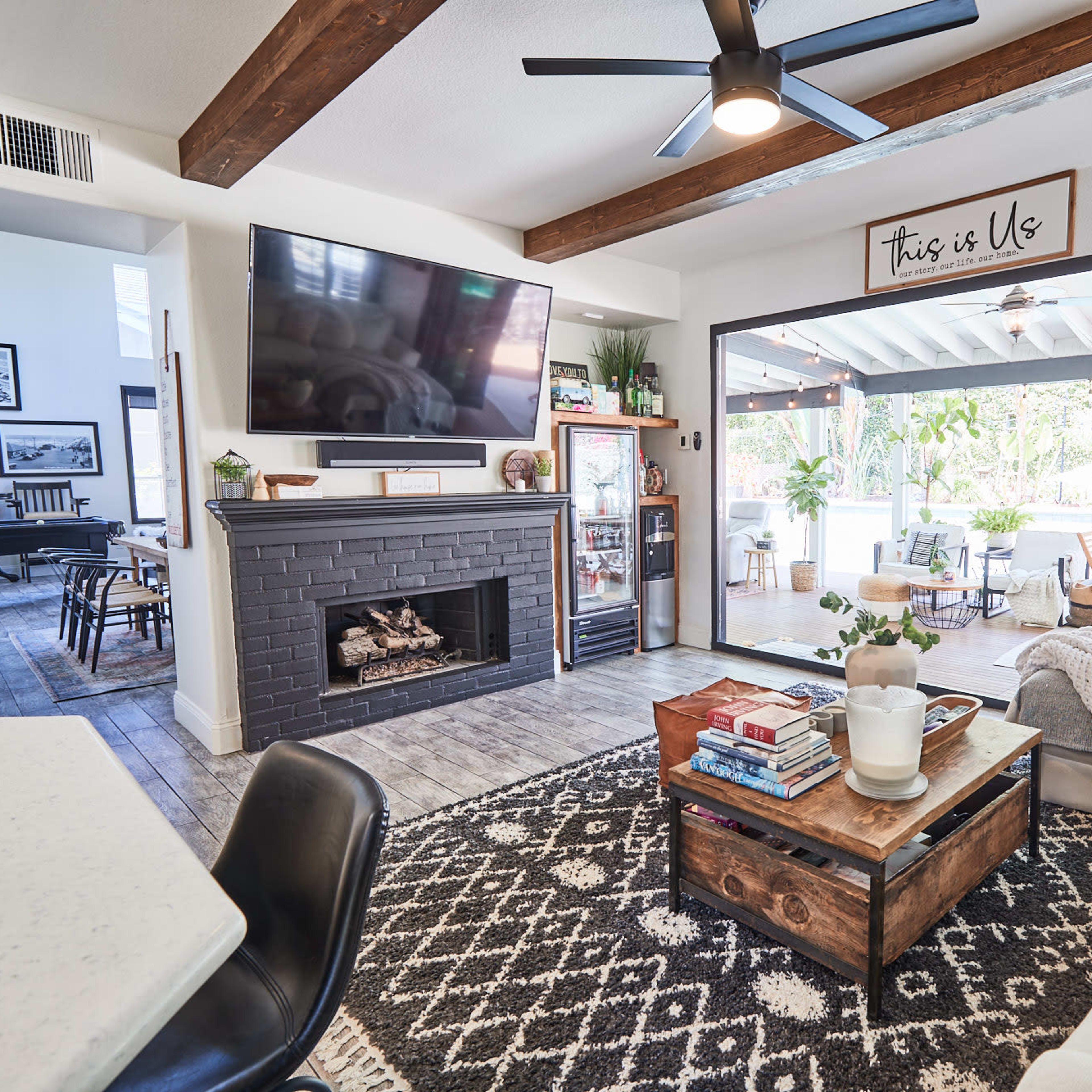 A living room features a modern fireplace, a large television, and a coffee table, with a view of an outdoor seating area through open glass doors.