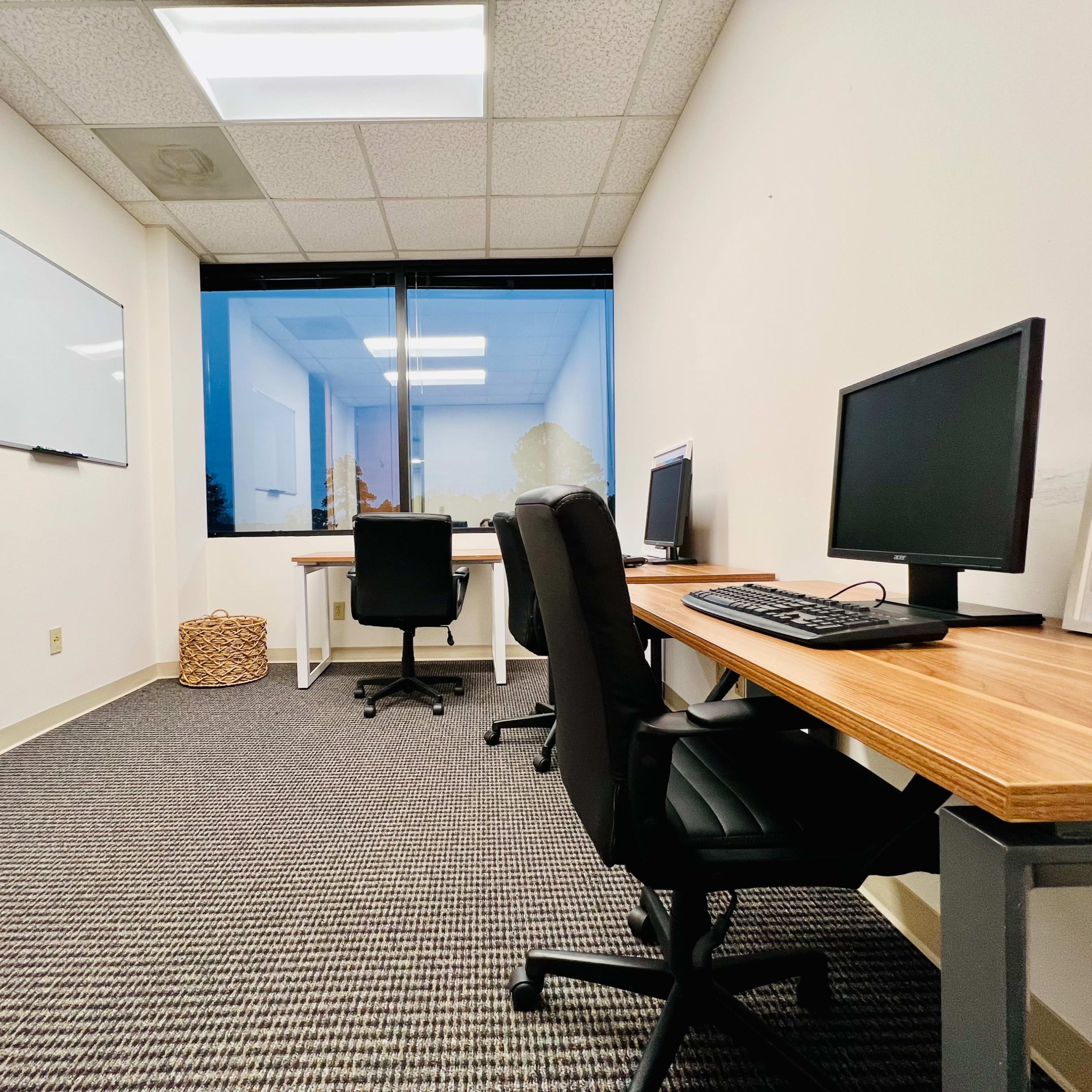 A sparsely furnished office space features three chairs and two computer desks, with a whiteboard on the wall and a large window displaying an outdoor view.