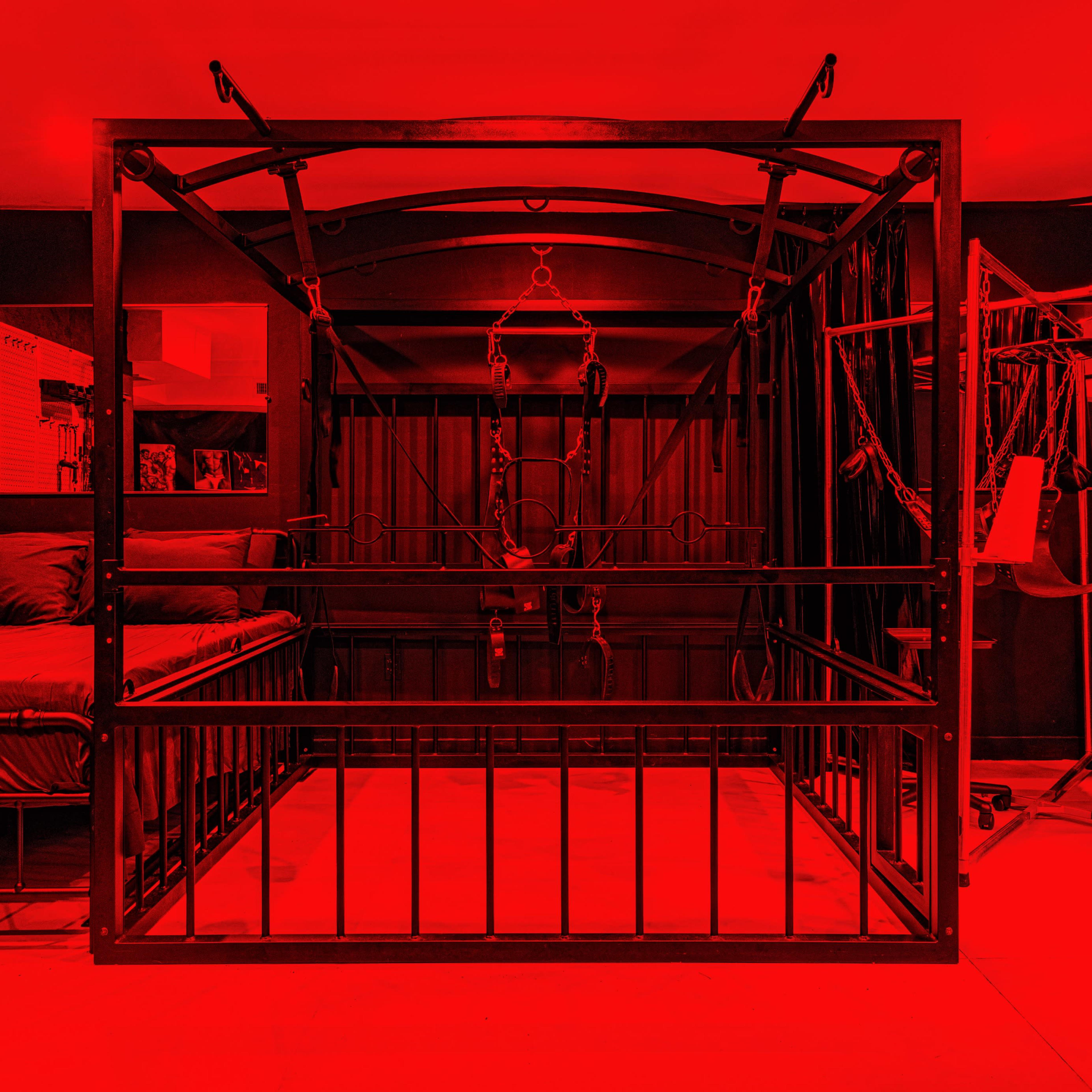 A red-lit room featuring a metal-frame bed with a surrounding bondage frame equipped with restraints.