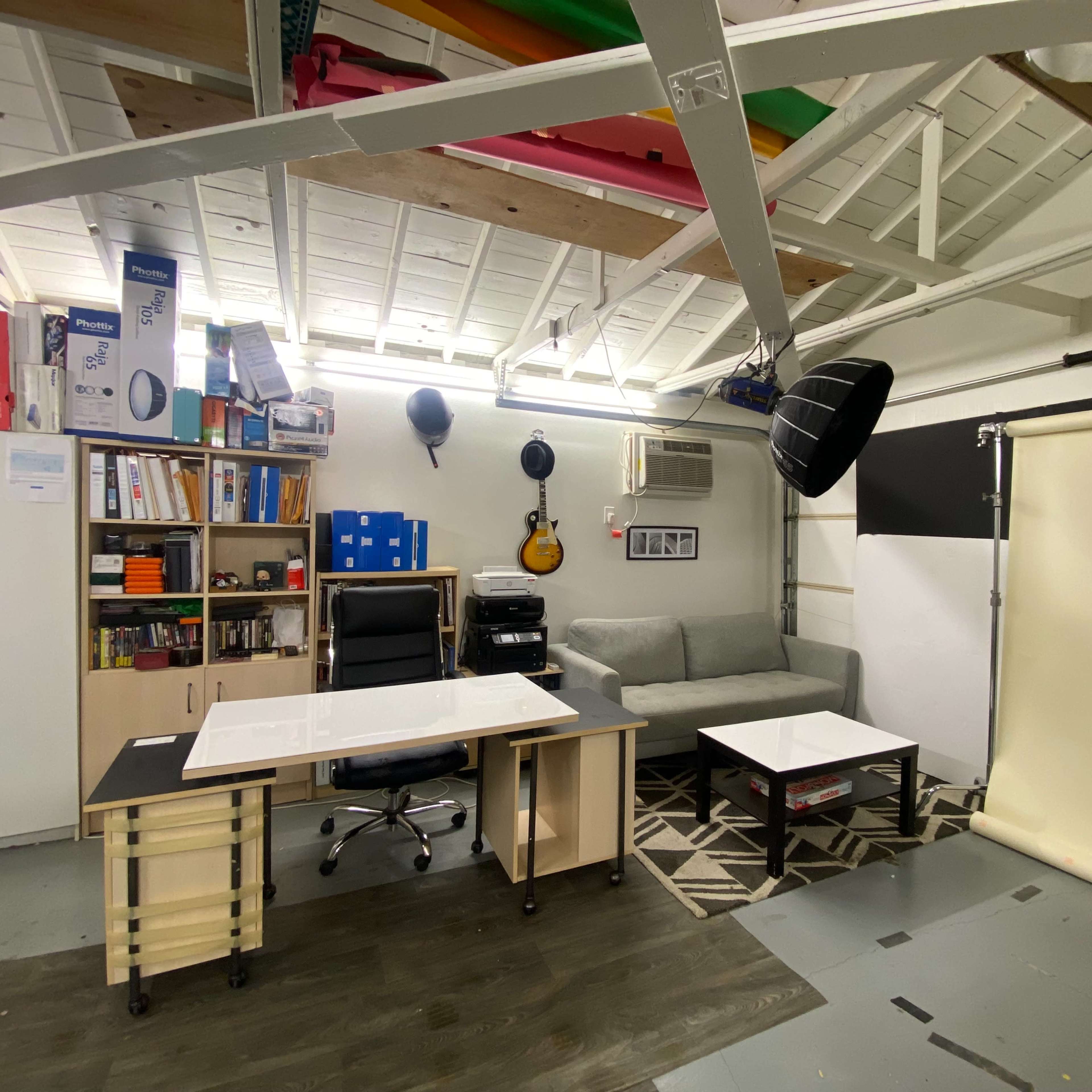 A workspace with a desk, office chair, a gray couch, a coffee table, and various storage items on shelves in a well-lit room.