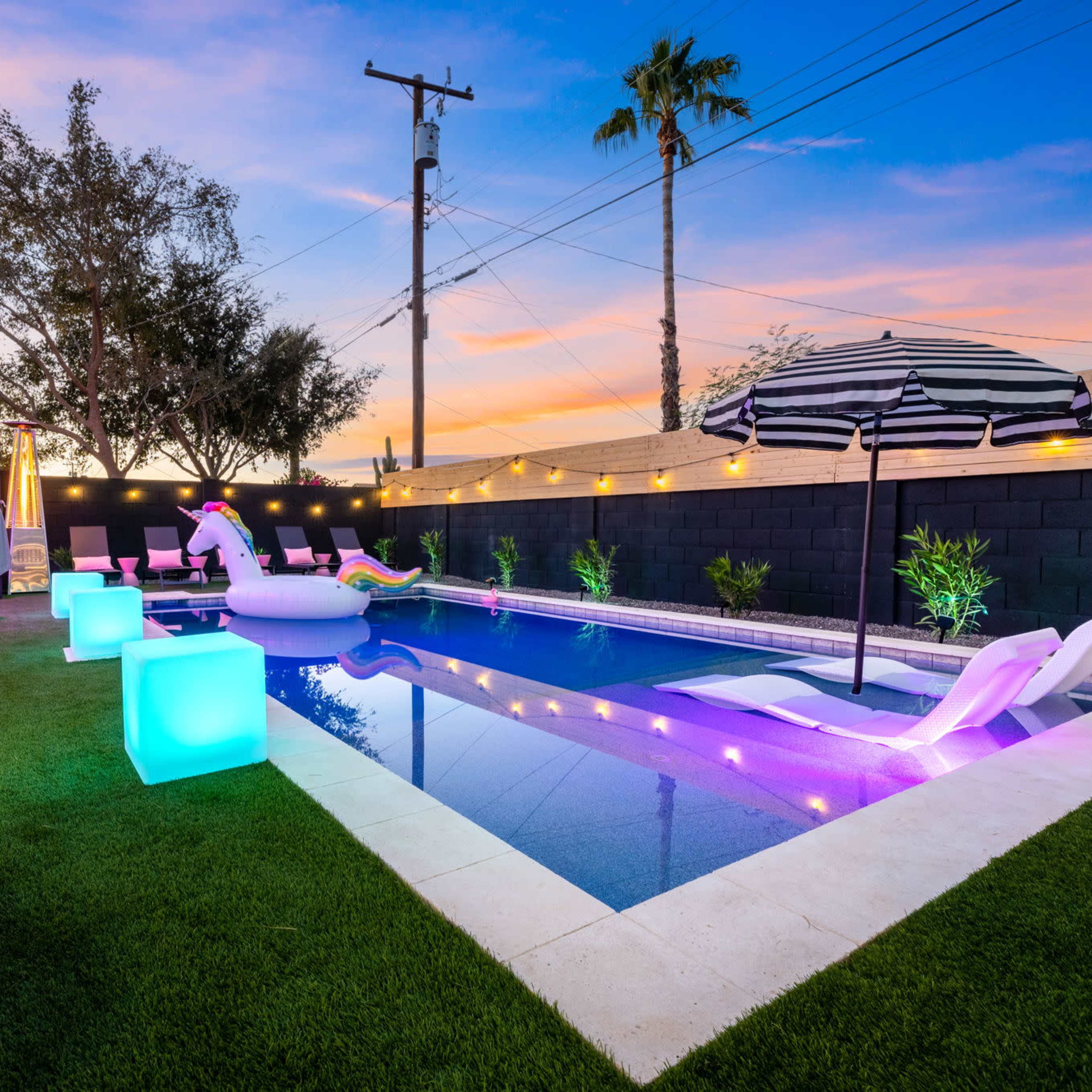 A backyard pool area with colorful lounge chairs, a large inflatable unicorn, and ambient lighting set against a sunset sky.