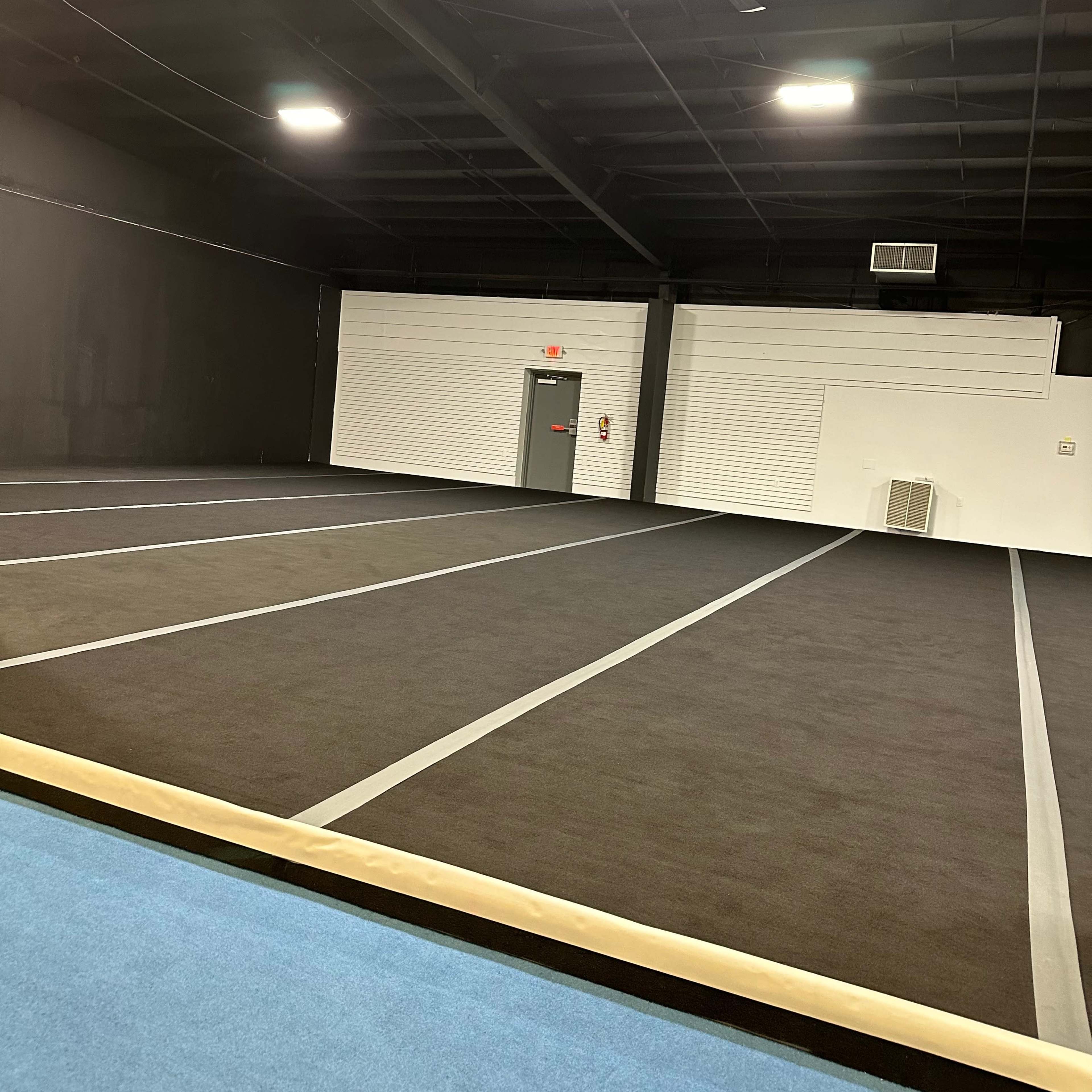 An indoor gymnasium features a large black matted area with a blue edge and a single gray door in the background.