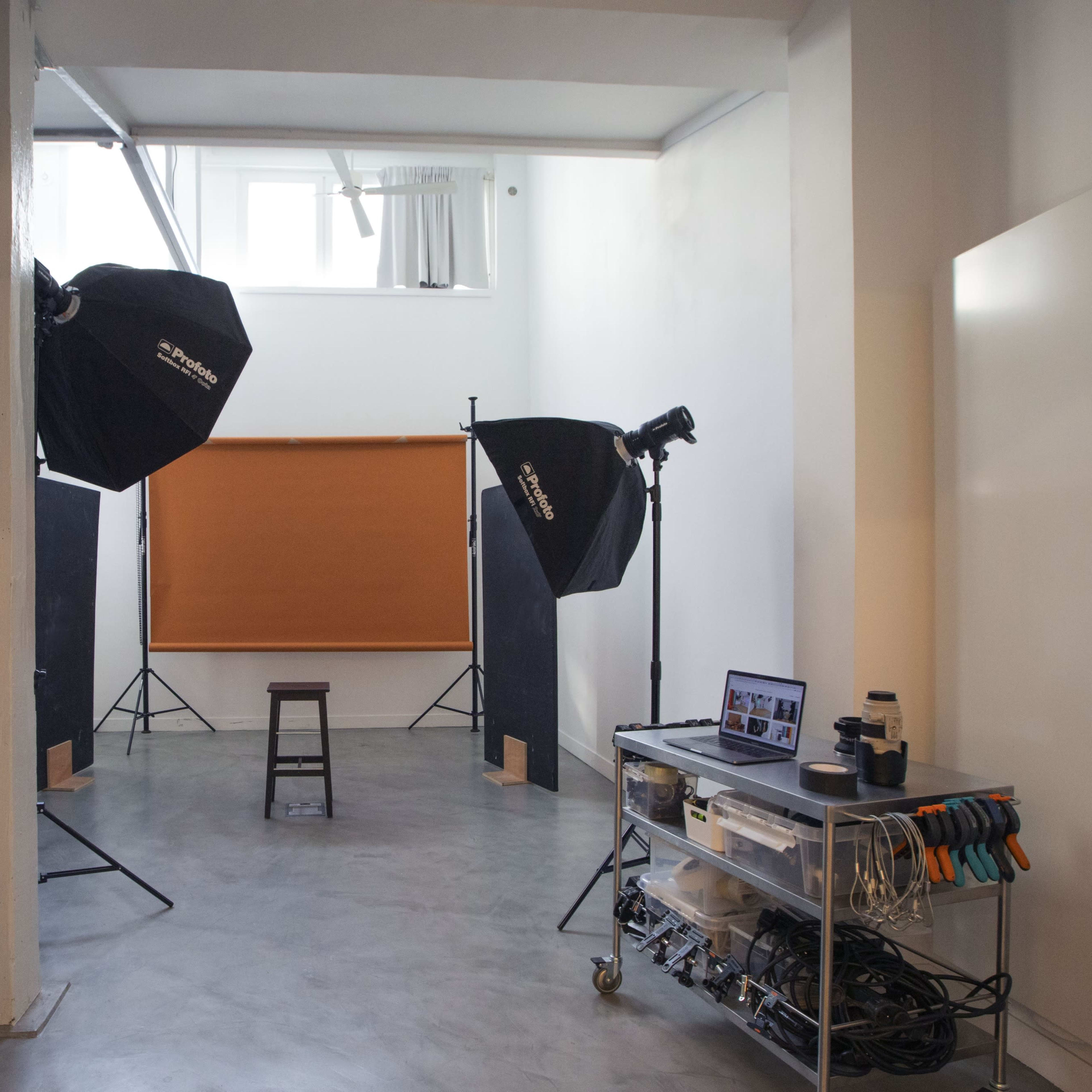 A photography studio contains two large softboxes, a backdrop, a stool, and various equipment on a rolling cart.