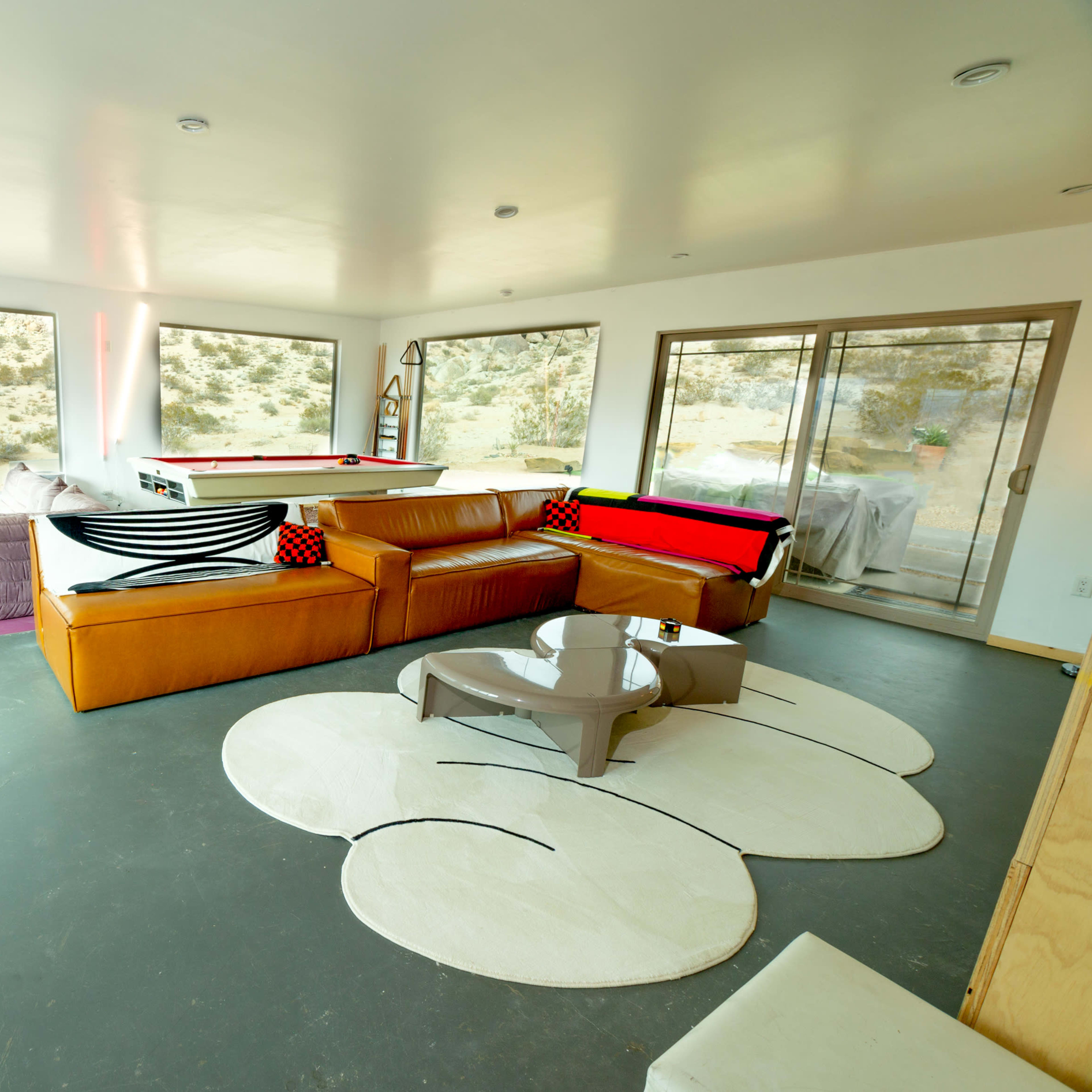 The room features large windows, a modern sectional sofa, a coffee table, and a pool table, all set against a backdrop of rocky terrain outside.