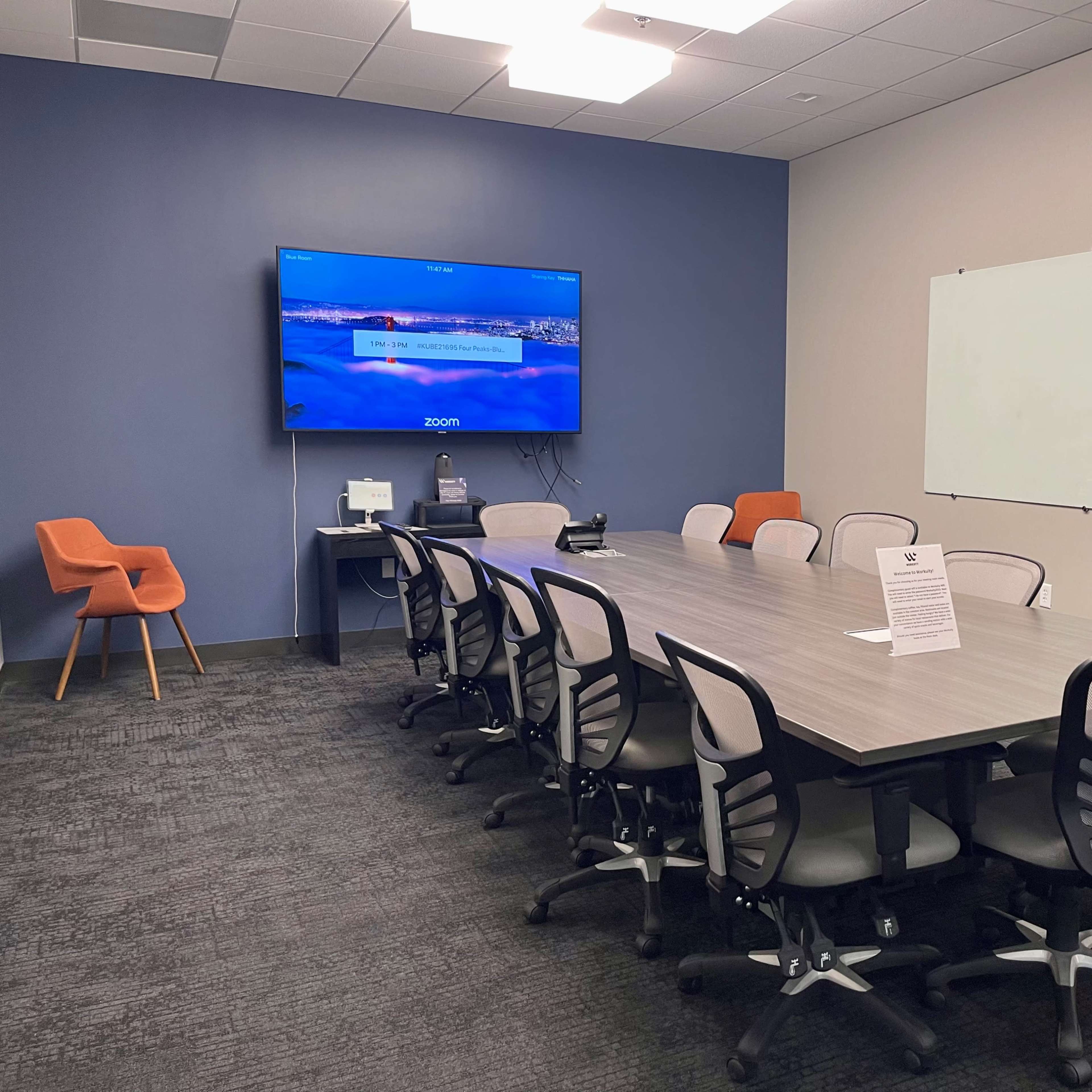 A conference room is set up with a long table surrounded by chairs, a large screen displaying a Zoom interface, and a whiteboard on the wall.