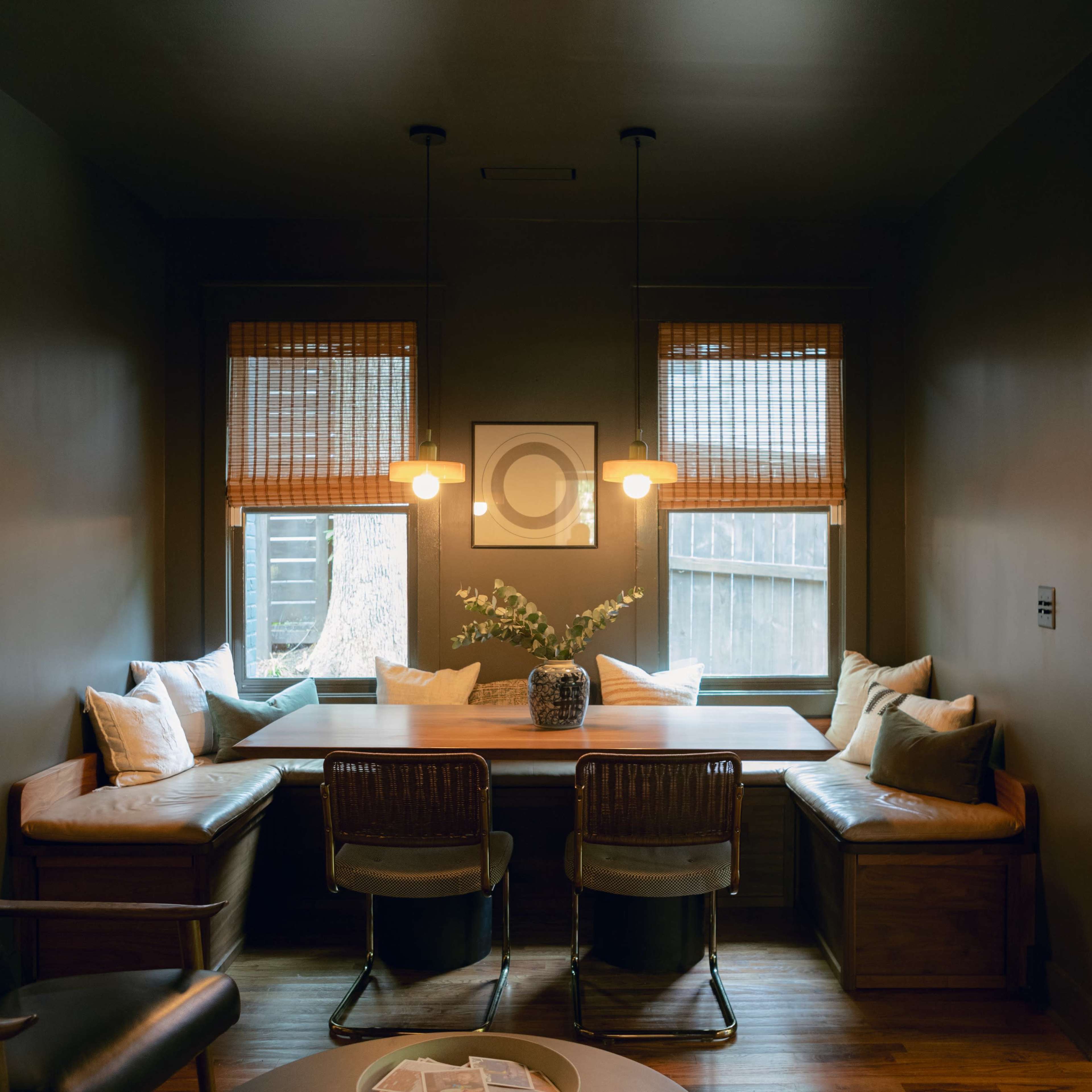 A cozy dining nook features a wooden bench surrounded by large windows, adorned with bamboo shades and a central round table with two metal chairs.