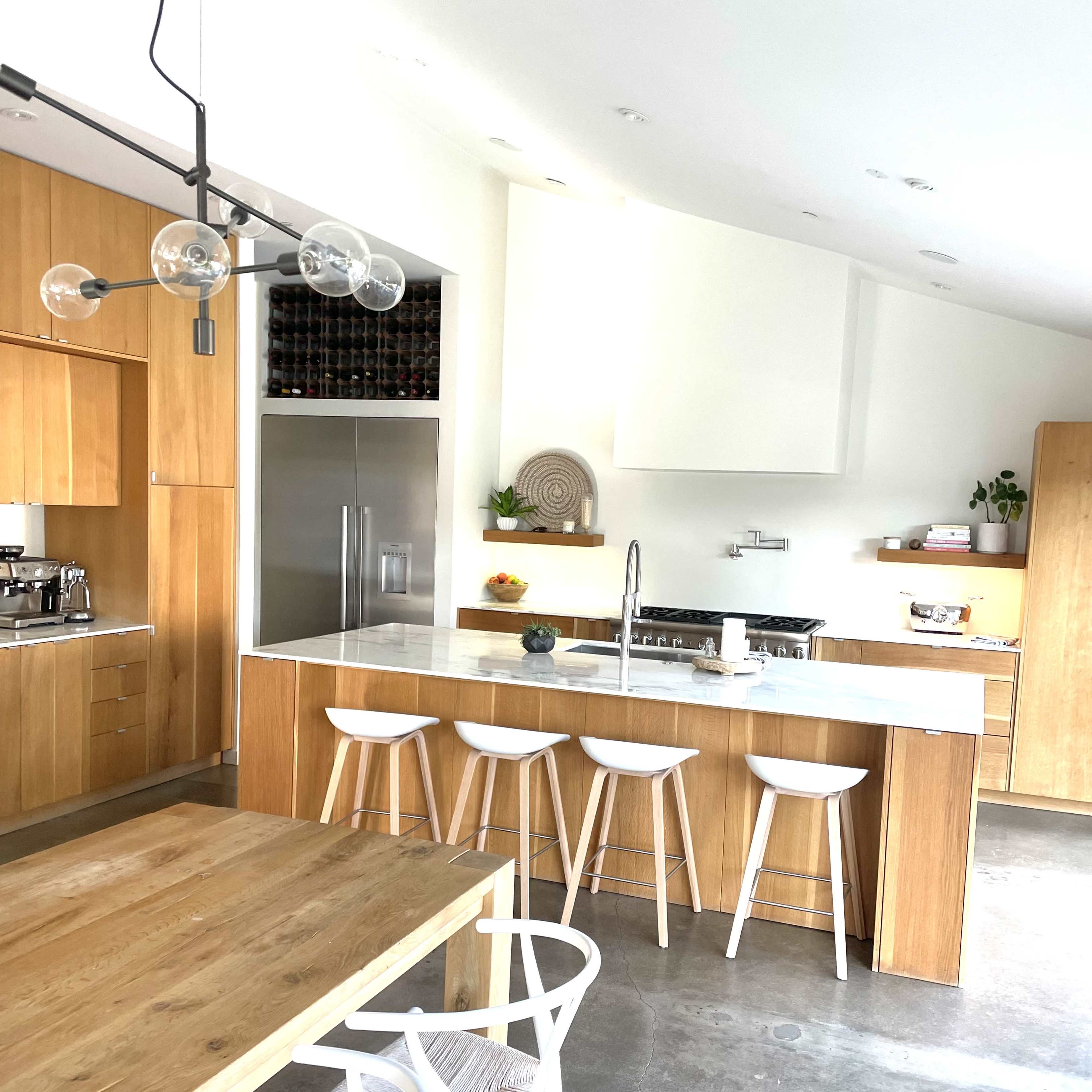 A modern kitchen with wooden cabinetry, a large island with white bar stools, and a dining table in the foreground.
