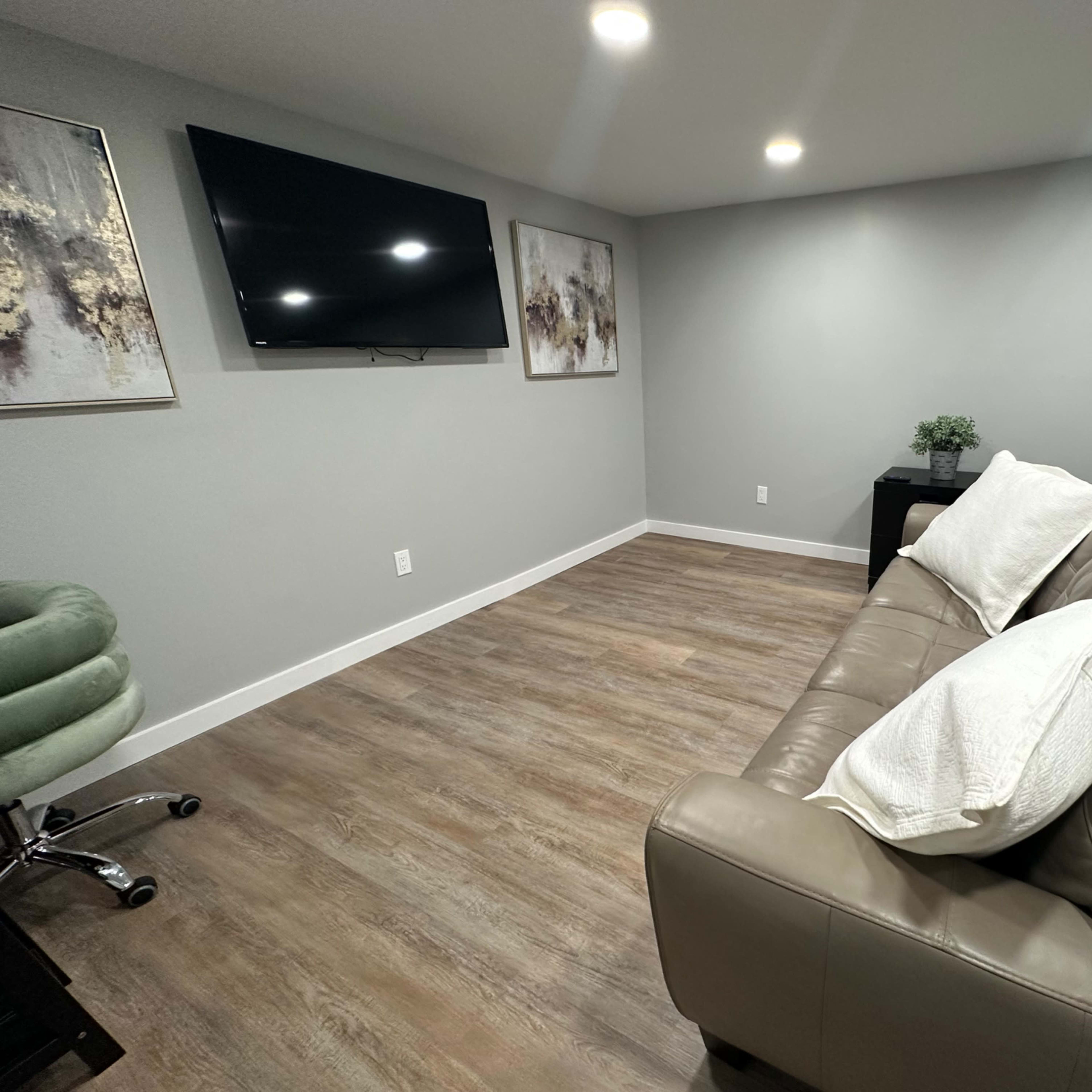 A modern, minimalistic living space with a light gray wall, a flat-screen TV mounted on the wall, a beige sofa with white cushions, and a small table beside it.