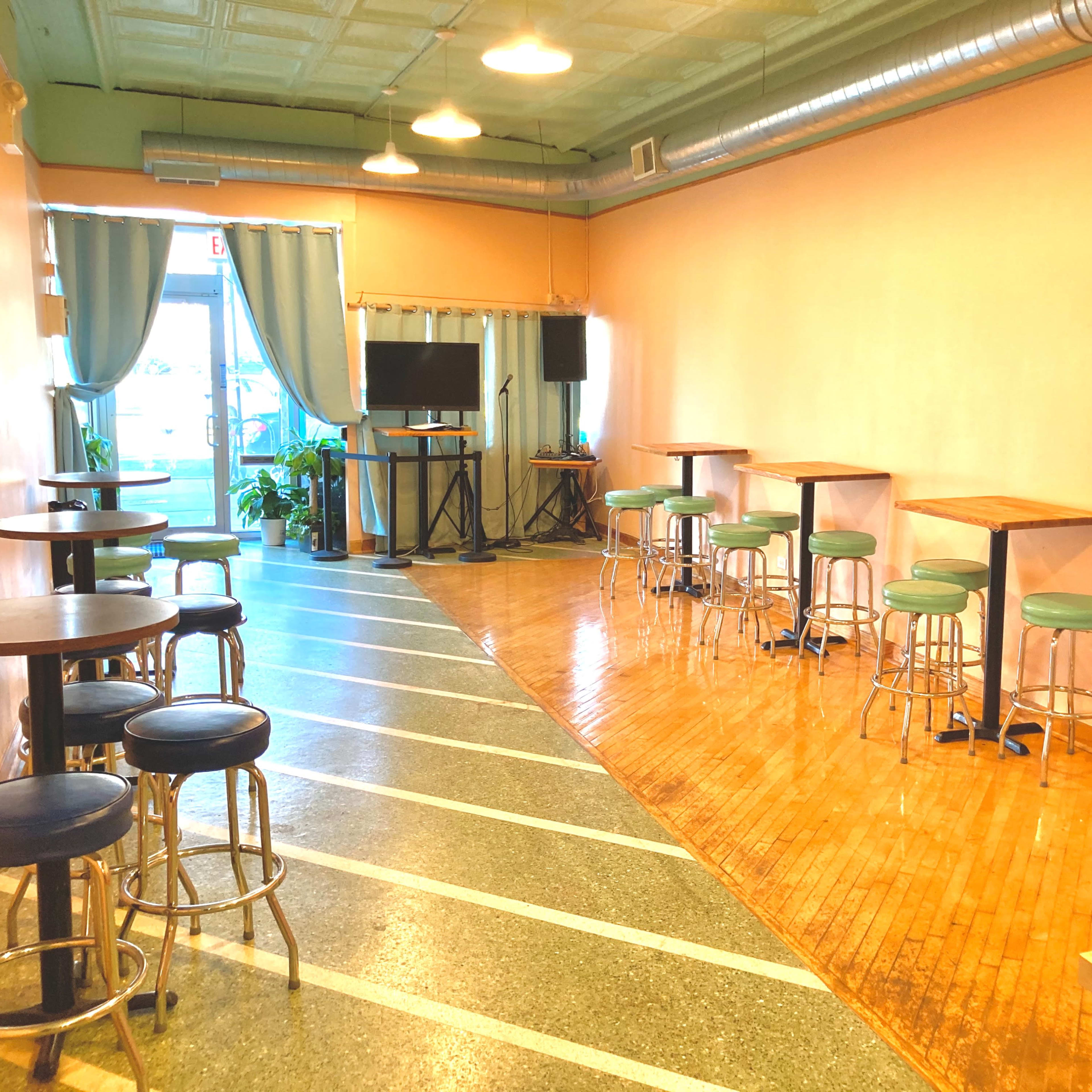 A spacious room with wooden floors, a row of tall tables and stools on one side, and large windows letting in natural light.