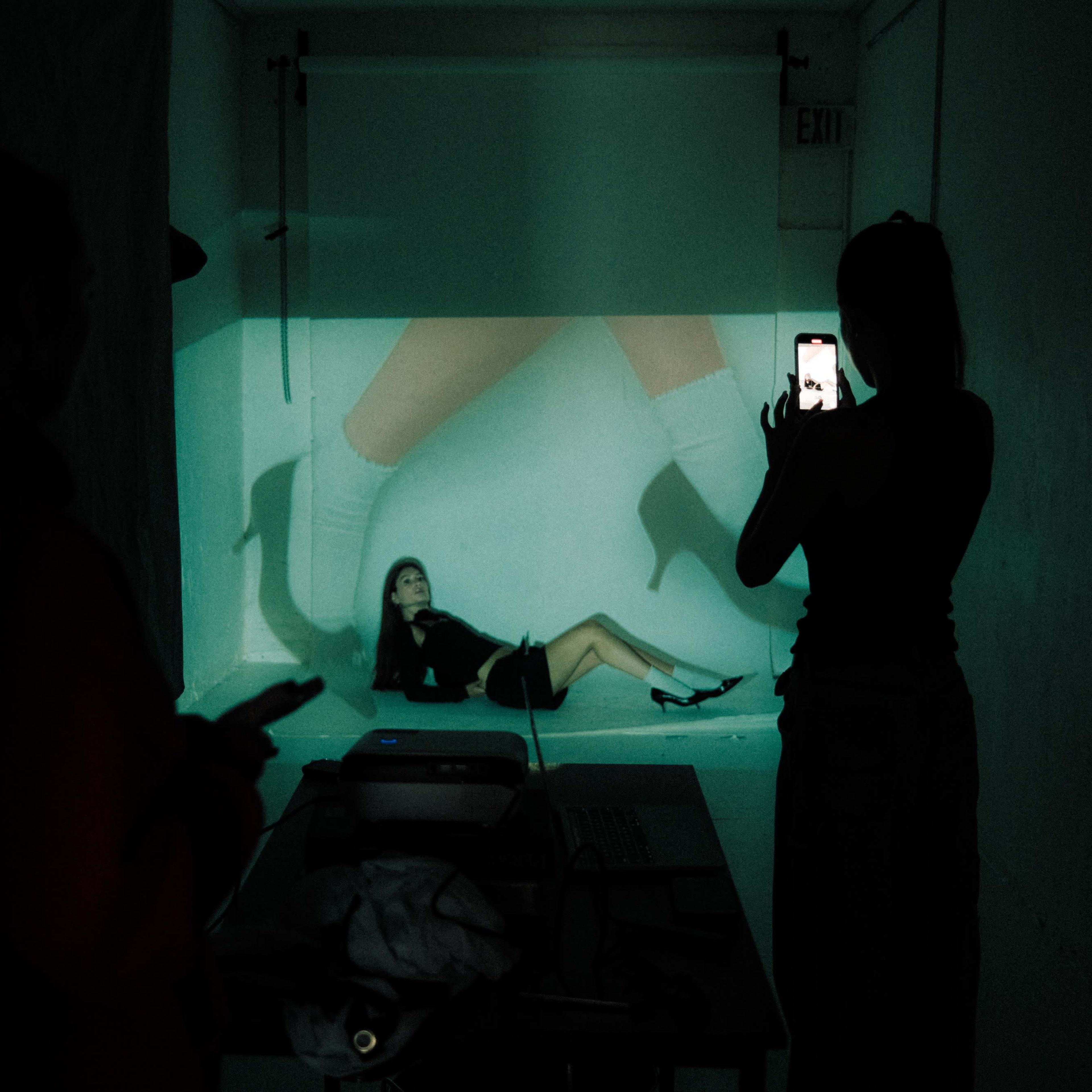 A woman poses on the ground in front of a projected image of legs, while another person takes a photo of her with a smartphone.