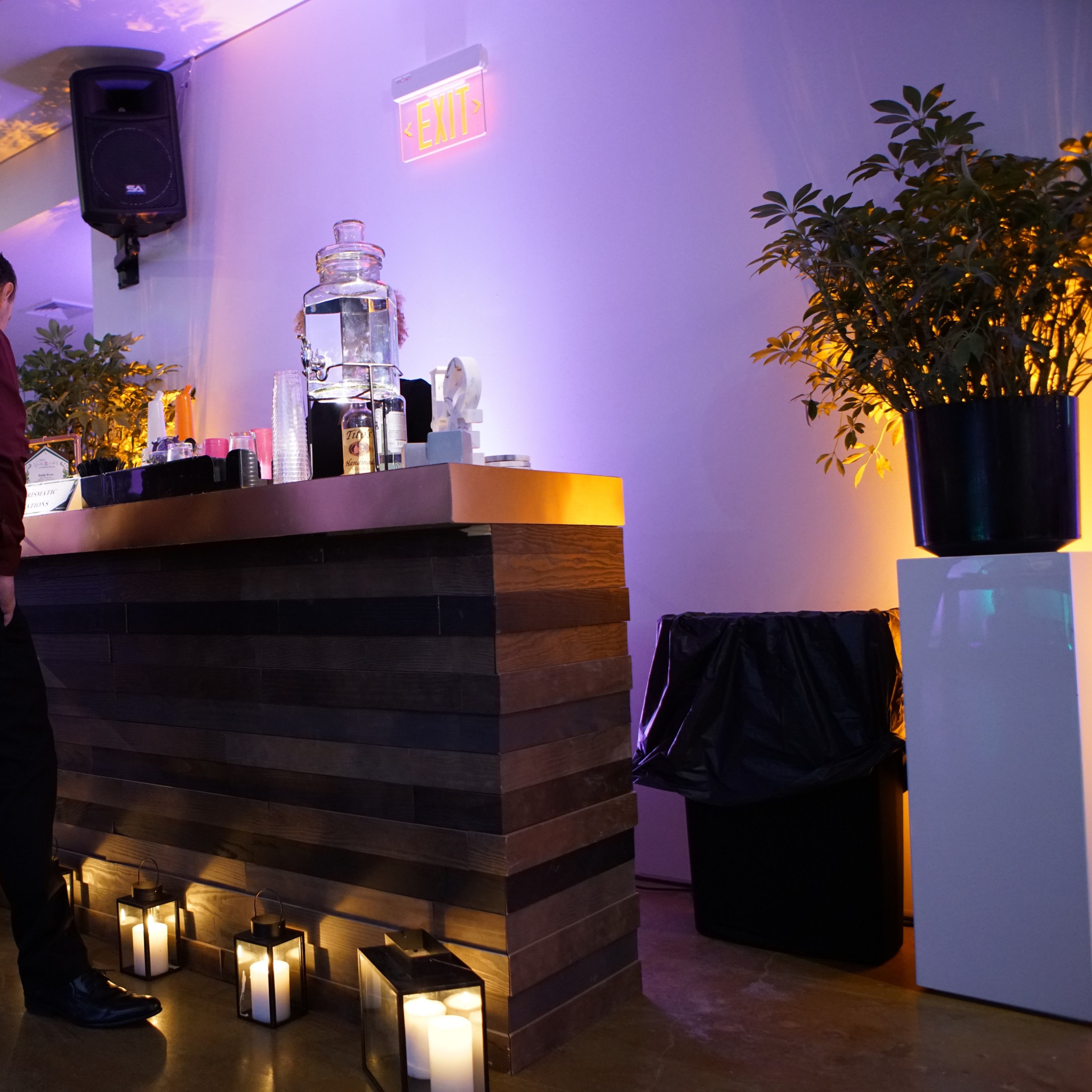 A man in a maroon shirt stands near a wooden bar illuminated by soft lighting, with decorative plants and candles arranged along the front.