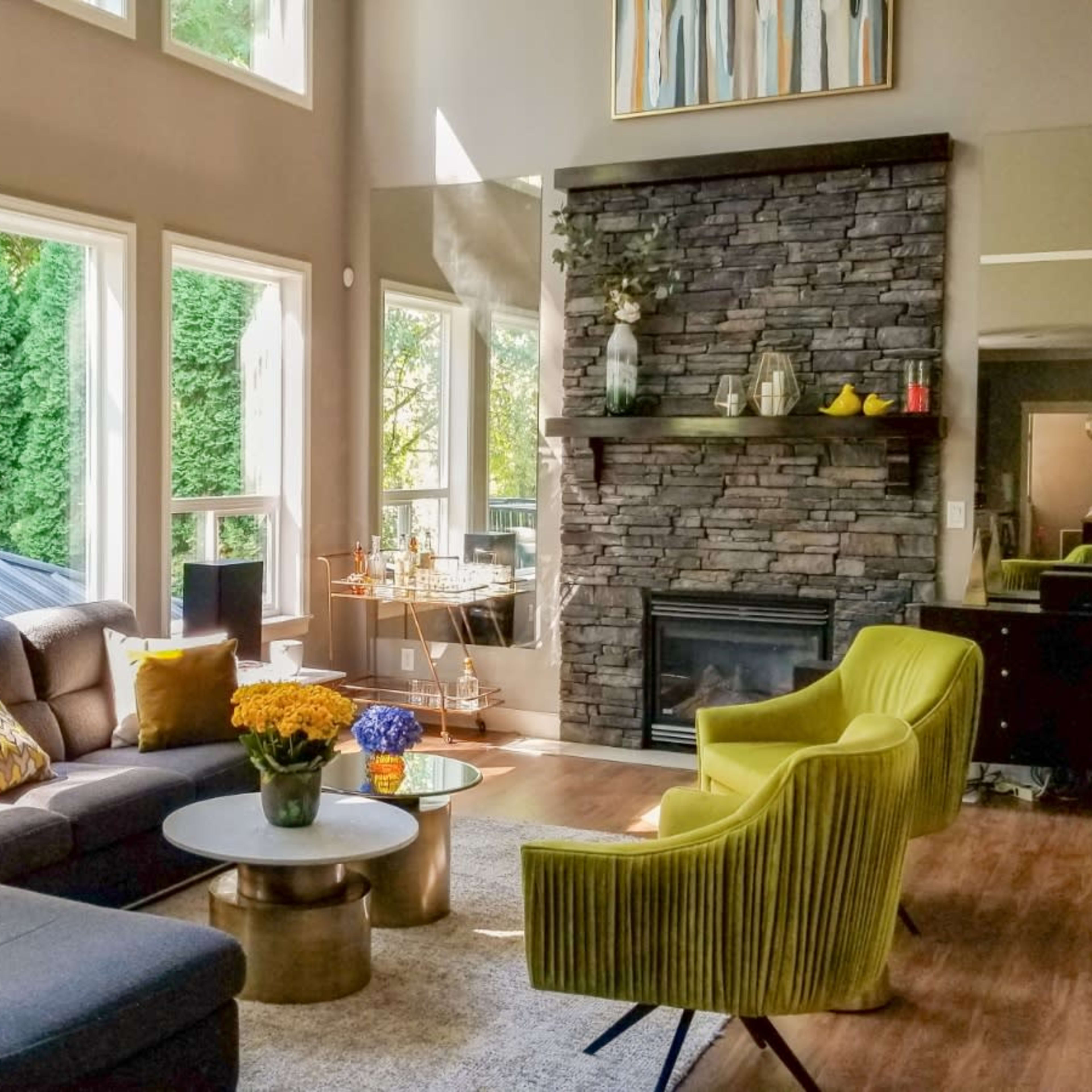 A modern living room with large windows, a stone fireplace, a circular coffee table, and bright yellow accent chairs.