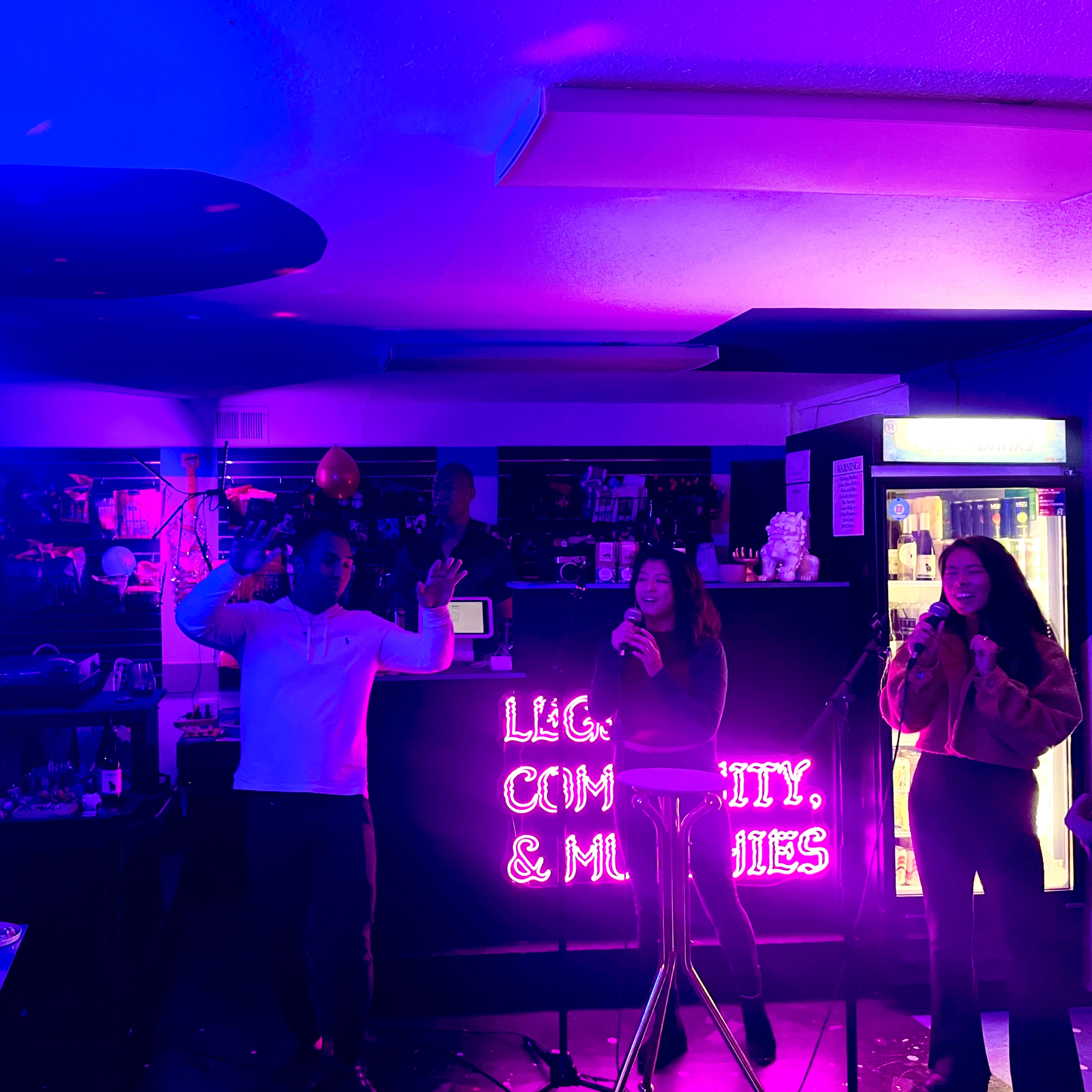 Two individuals sing karaoke while a third person stands behind a bar in a brightly lit space adorned with purple and pink hues.