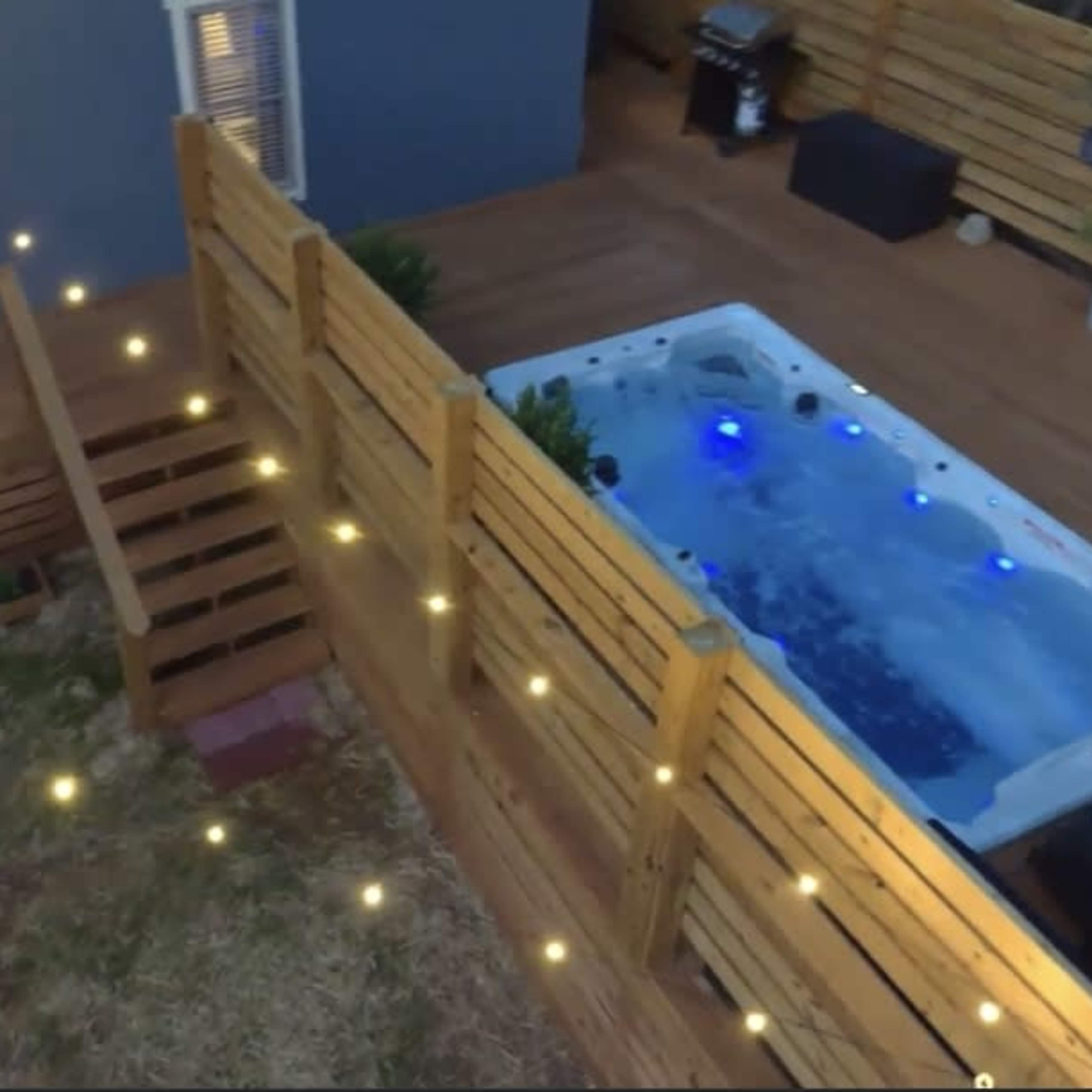 A backyard with a fenced area featuring a hot tub with blue lights, a wooden deck, and outdoor furniture.
